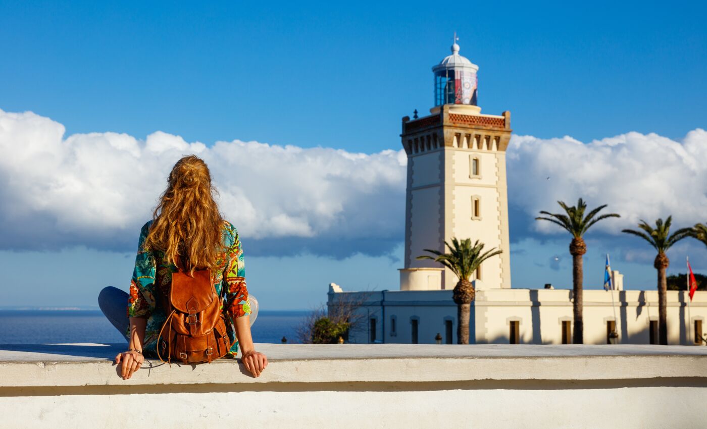  A person sits in contemplation, facing a lighthouse and palm trees against a backdrop of a deep blue sea and fluffy white clouds.