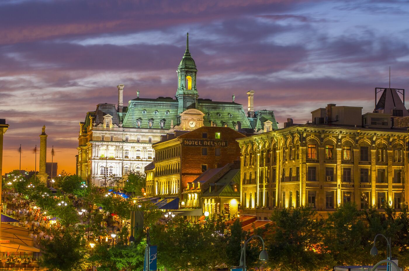 Twilight descends on a lively city, highlighting historical buildings with glowing lights against a dramatic sky.