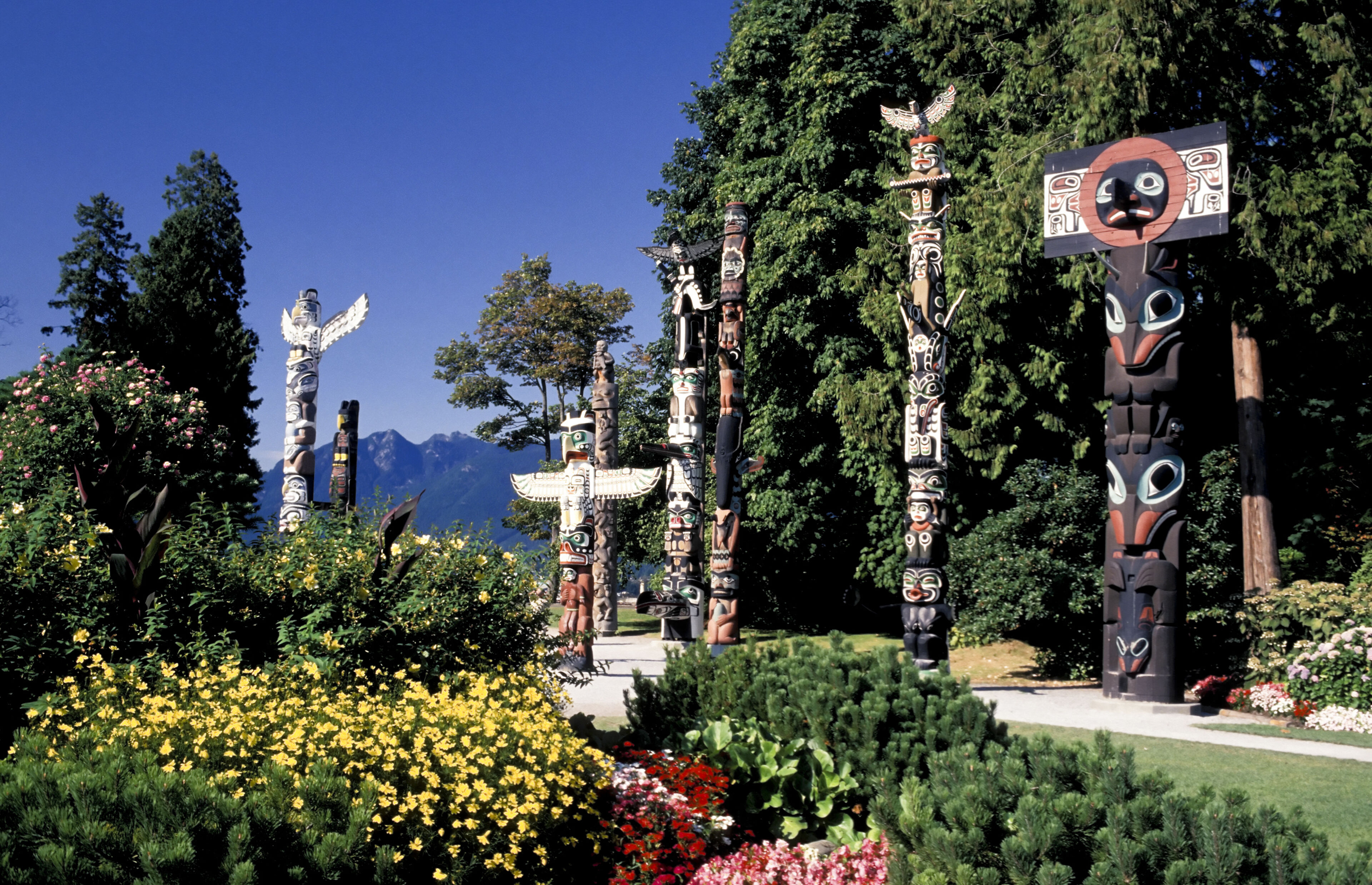 A row of intricately carved totem poles stands majestically in a blooming garden with mountain scenery in the background.