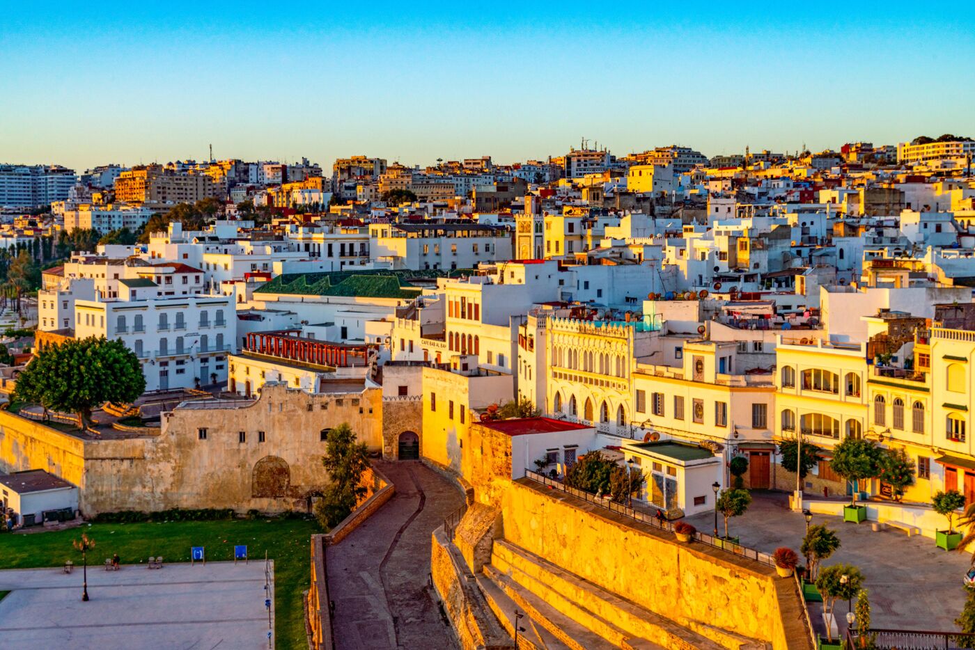 The image features a city with white buildings, a brick wall, and a street with cars and people. It is a sunny day, and the scene is captured from a high vantage point, giving a panoramic view of the city.