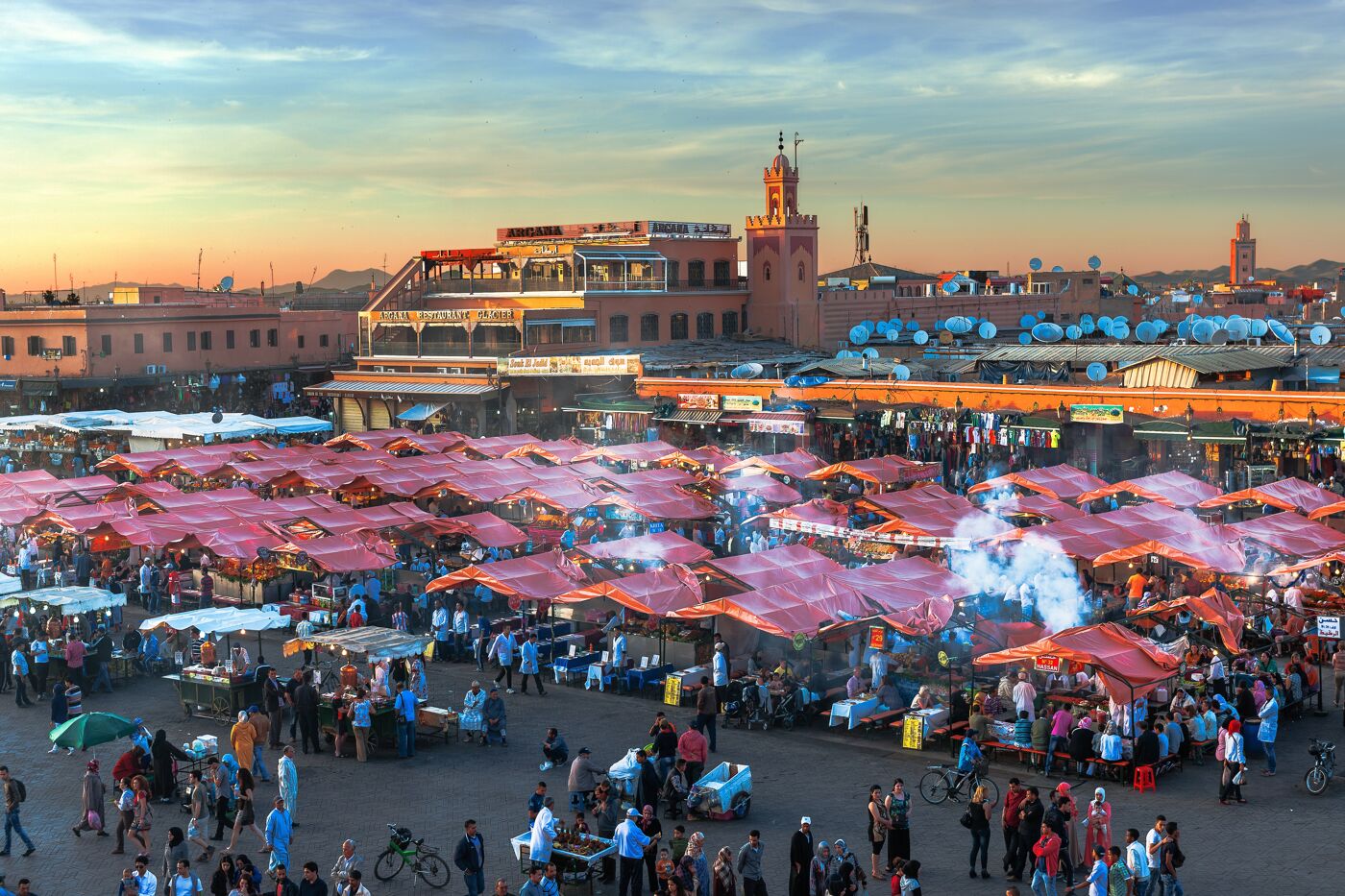 A lively open-air market teeming with activity, colorful stalls, and smoky street food vendors against a sunset sky.