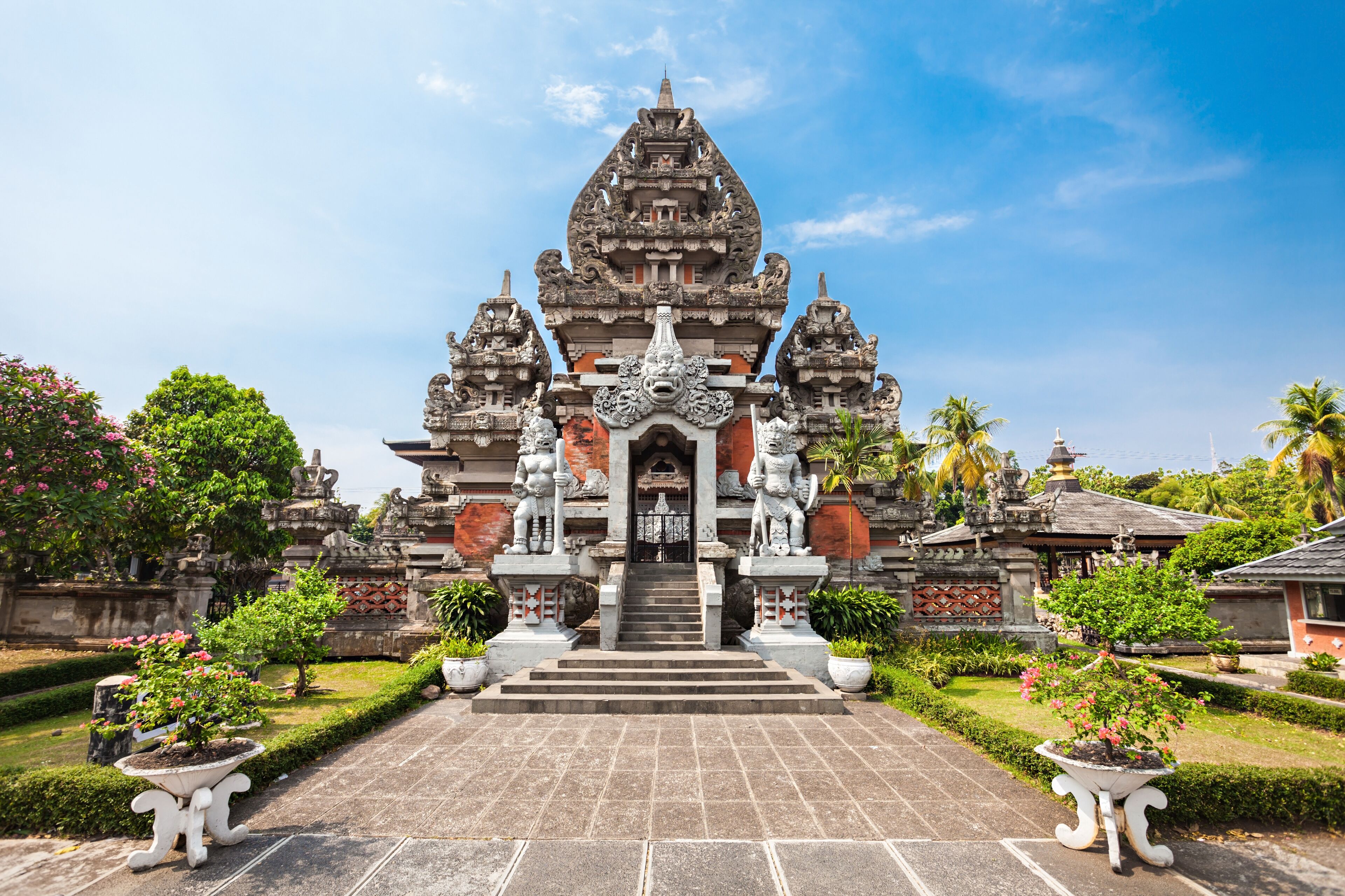 The entrance to a Balinese temple, featuring intricate stone carvings, guardian statues, and tropical foliage under a clear sky.