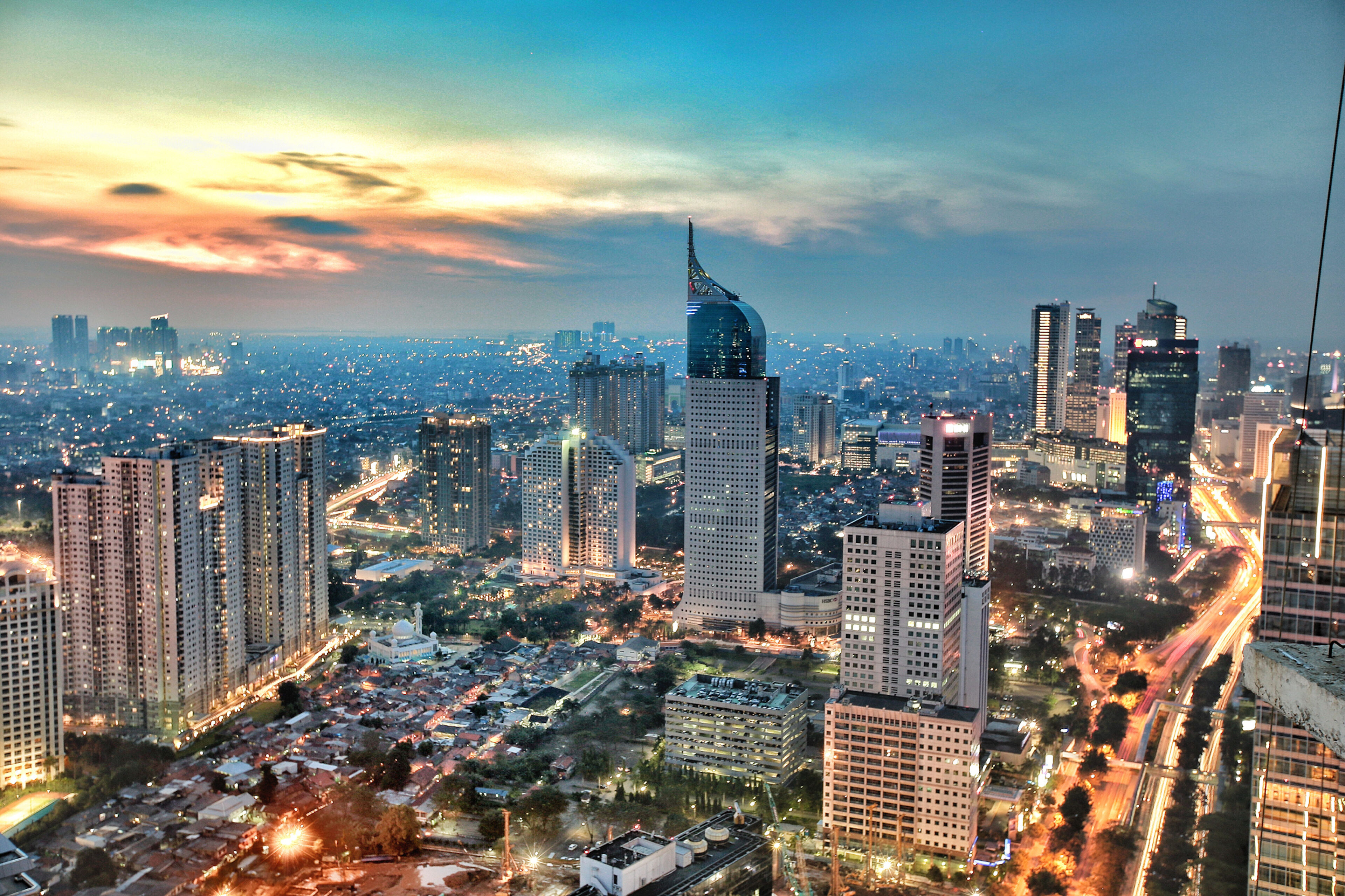 The skyline of Jakarta during twilight with illuminated skyscrapers and a vibrant sunset backdrop.