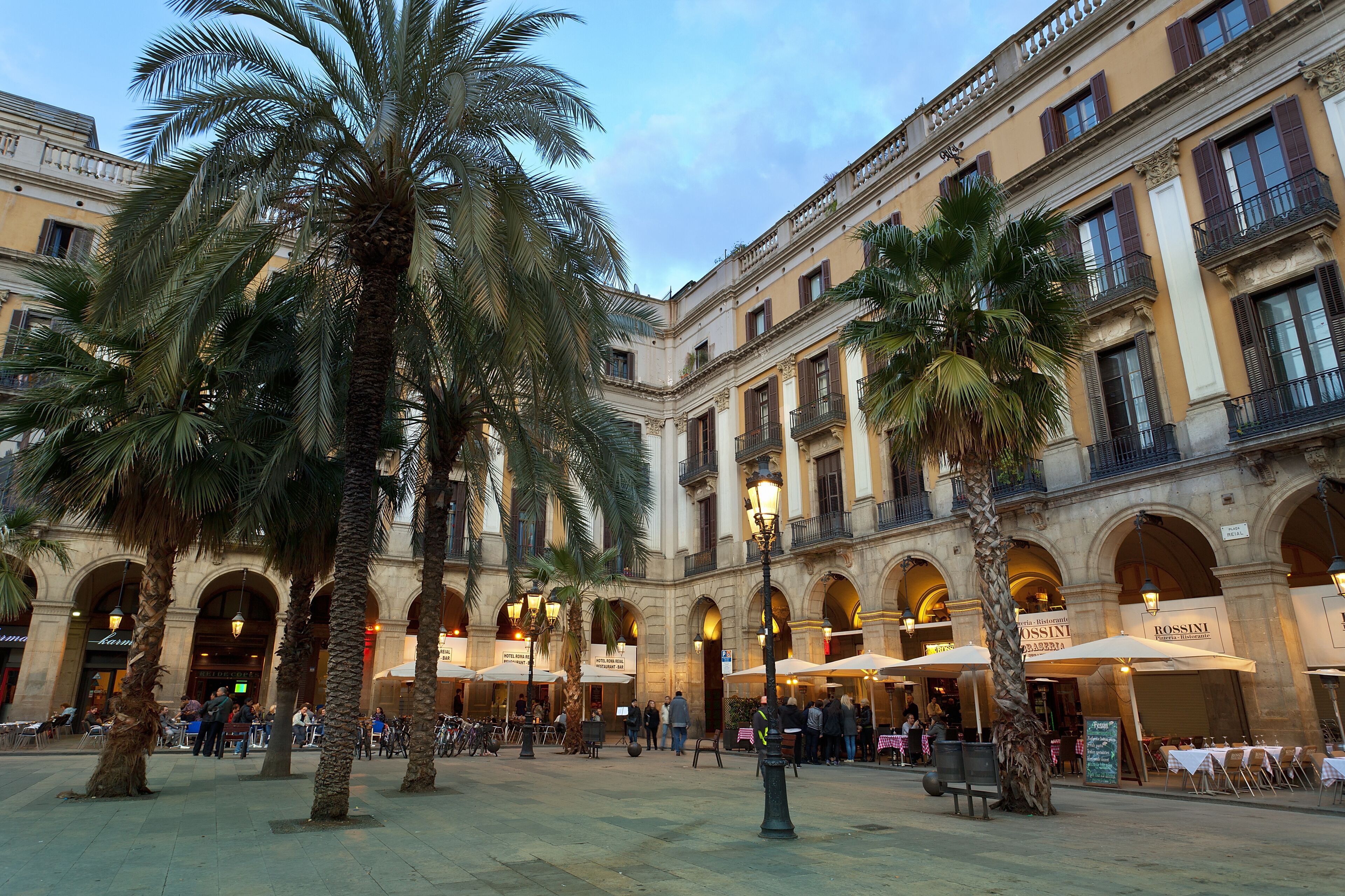 A bustling Mediterranean plaza with palm trees, outdoor cafes, and classic architecture at twilight, evoking a sense of relaxed urban life.