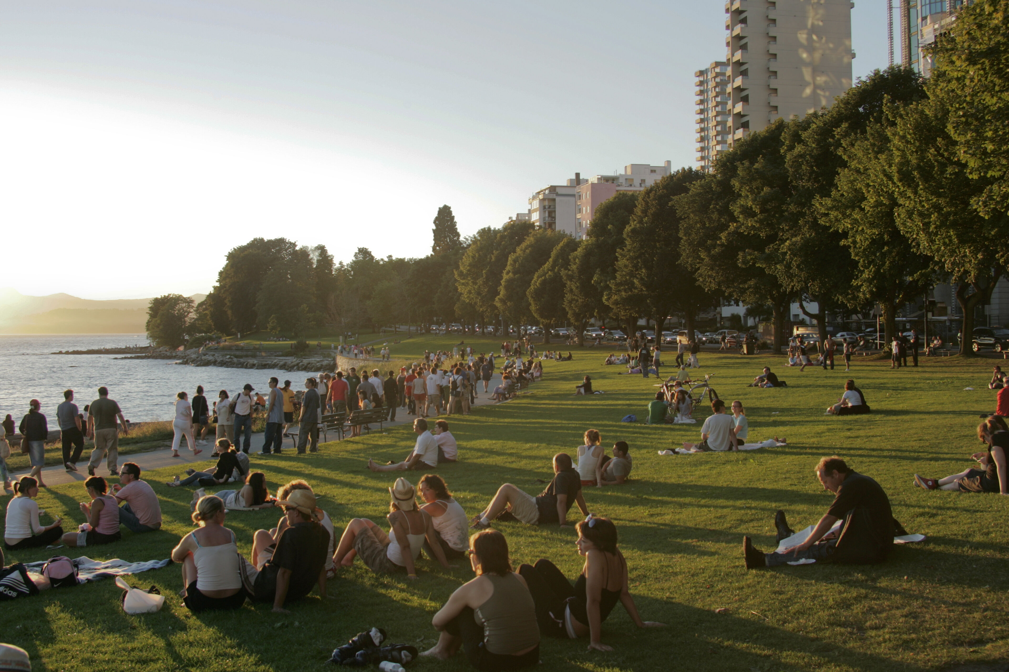 A serene sunset scene at an urban park with people lounging on the grass and walking along the waterfront, enjoying the warm evening light.