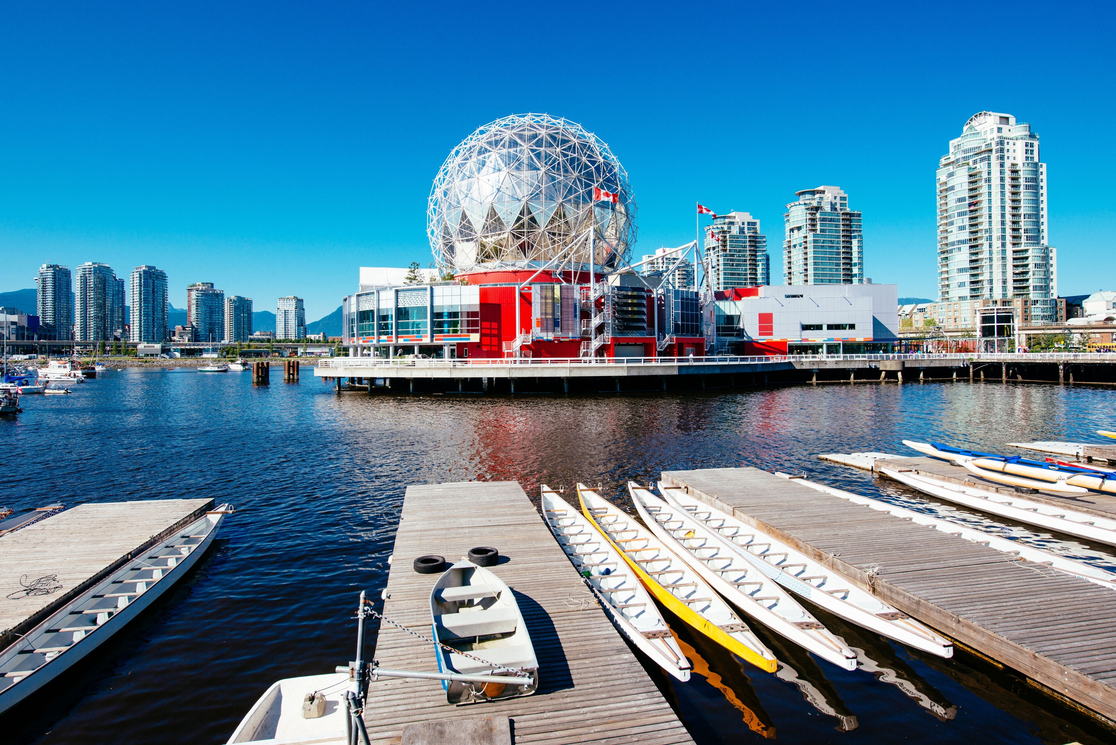 A geodesic dome structure stands by the waterfront, surrounded by modern buildings, with kayaks lined up on the dock.