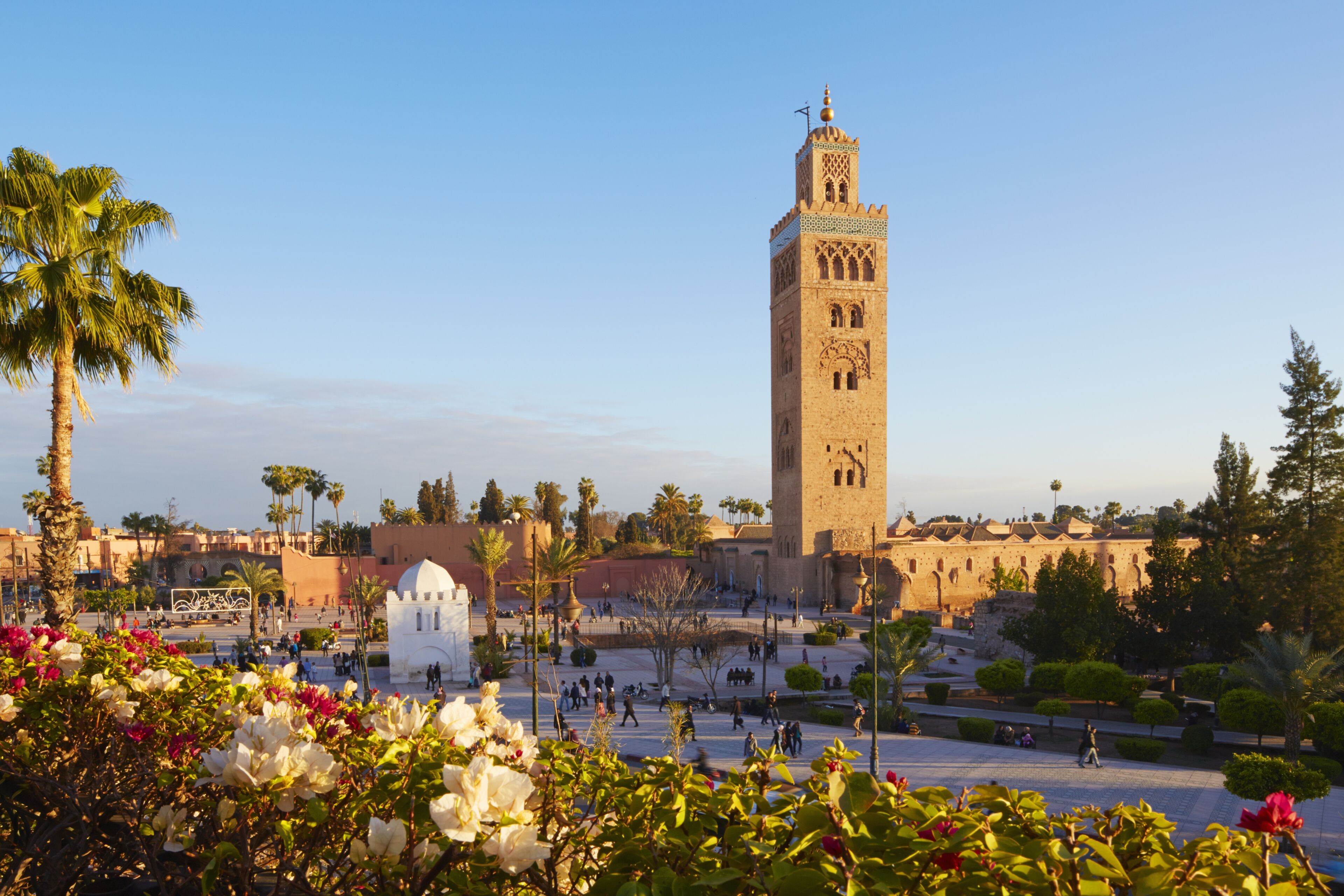 A towering minaret rises above a lively square surrounded by gardens and palms, under the soft light of late afternoon.