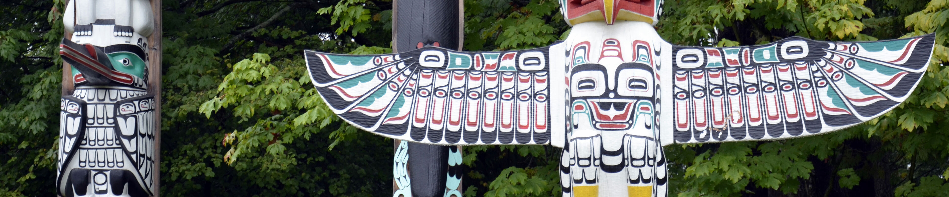 Colorful totem poles adorned with traditional indigenous carvings stand before a backdrop of dense green foliage.