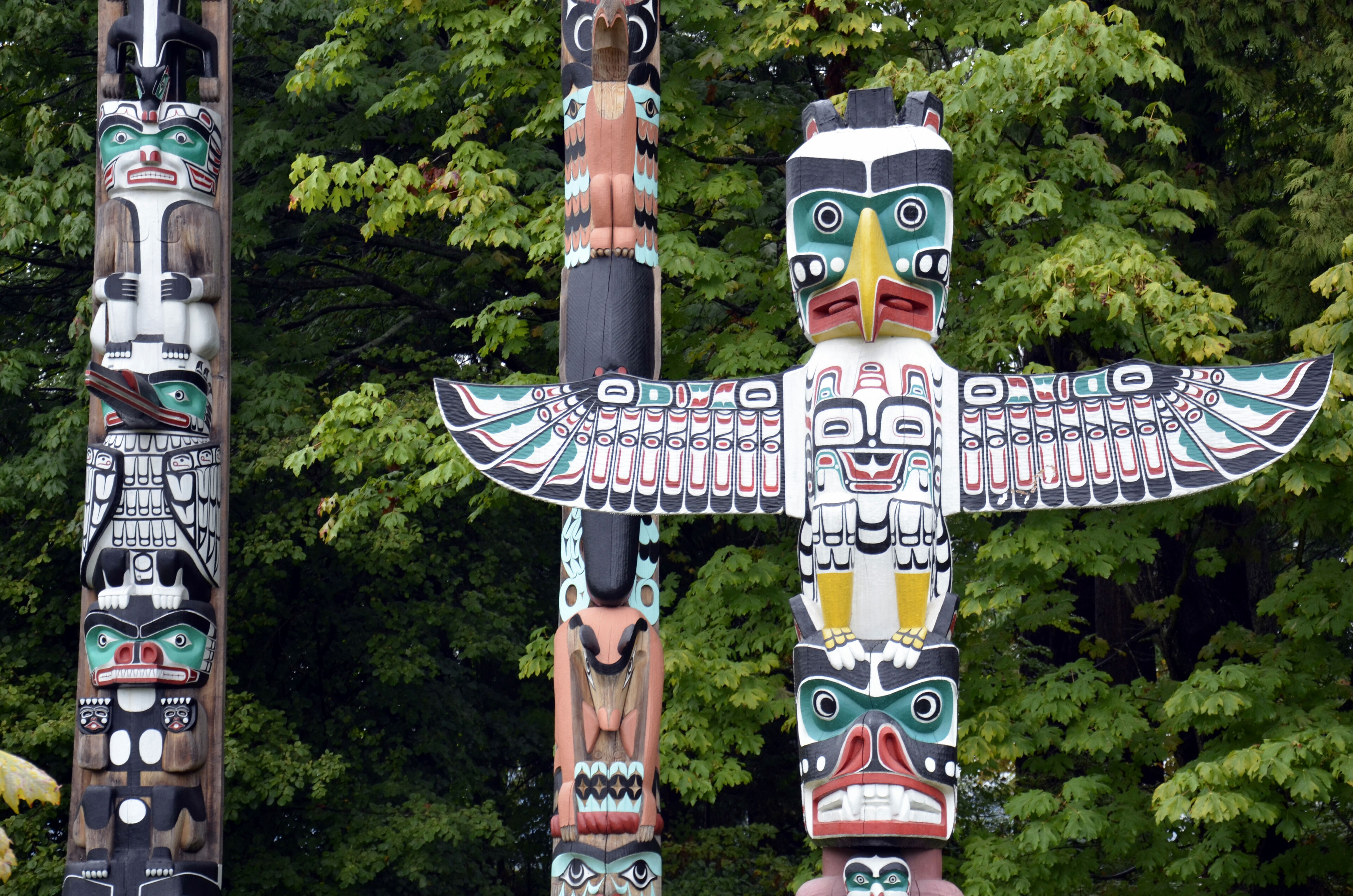 Colorful totem poles adorned with traditional indigenous carvings stand before a backdrop of dense green foliage.