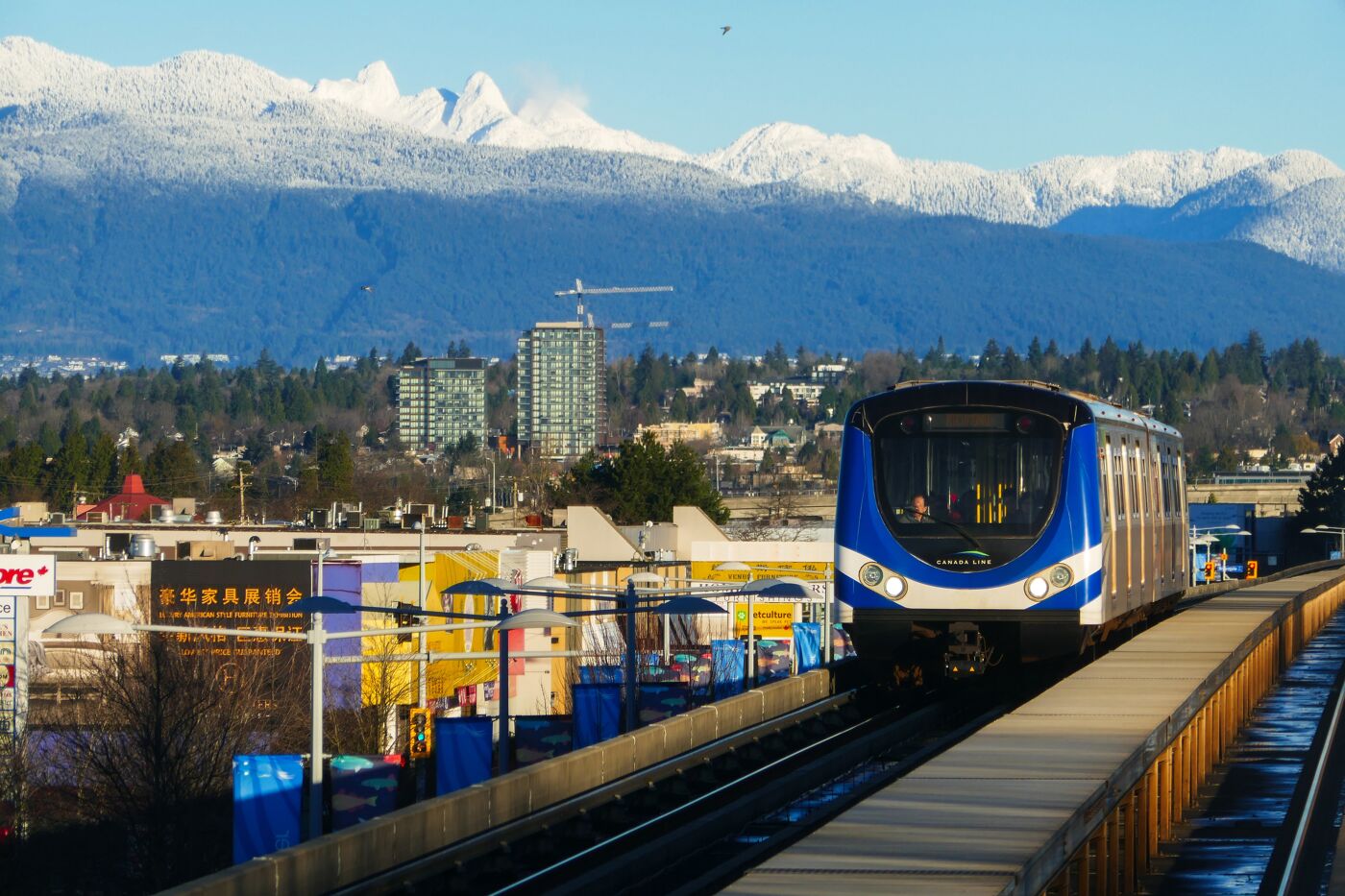 In winter, the Canada Line rapid transit on the elevated railway with city streets and buildings views and snow capped mountains as backdrop, between Richmond and Vancouver cities, in BC Canada.