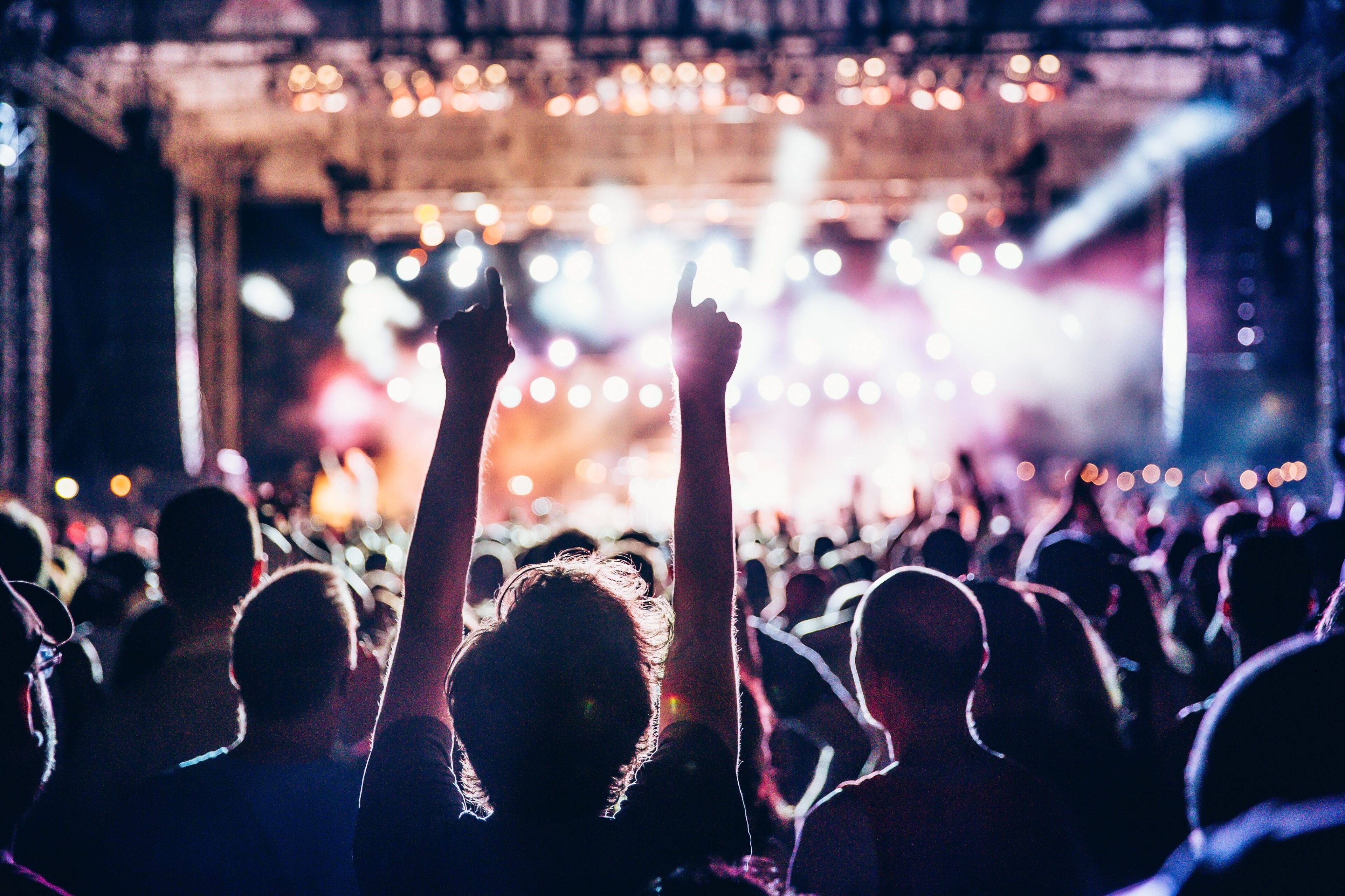 Silhouetted concert-goers raise hands against a vibrant stage lighting at an outdoor music festival at night.