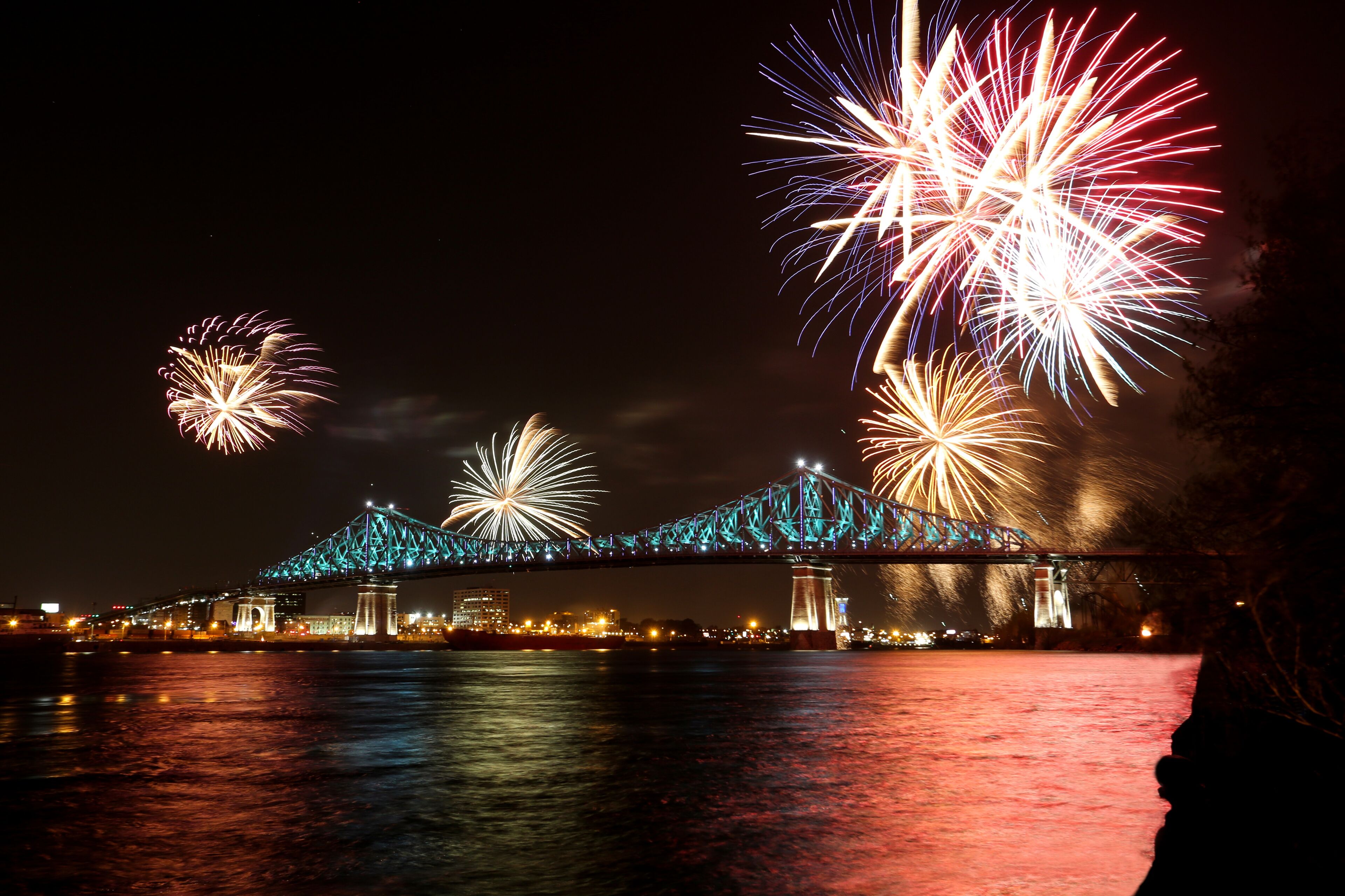 A spectacular fireworks show illuminates the sky above a brightly lit bridge spanning a river at night.