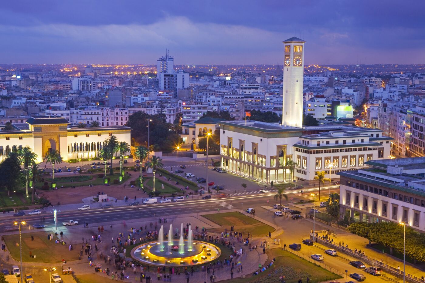 A vibrant city square at twilight with illuminated buildings, active fountains, and bustling crowds enjoying the evening.