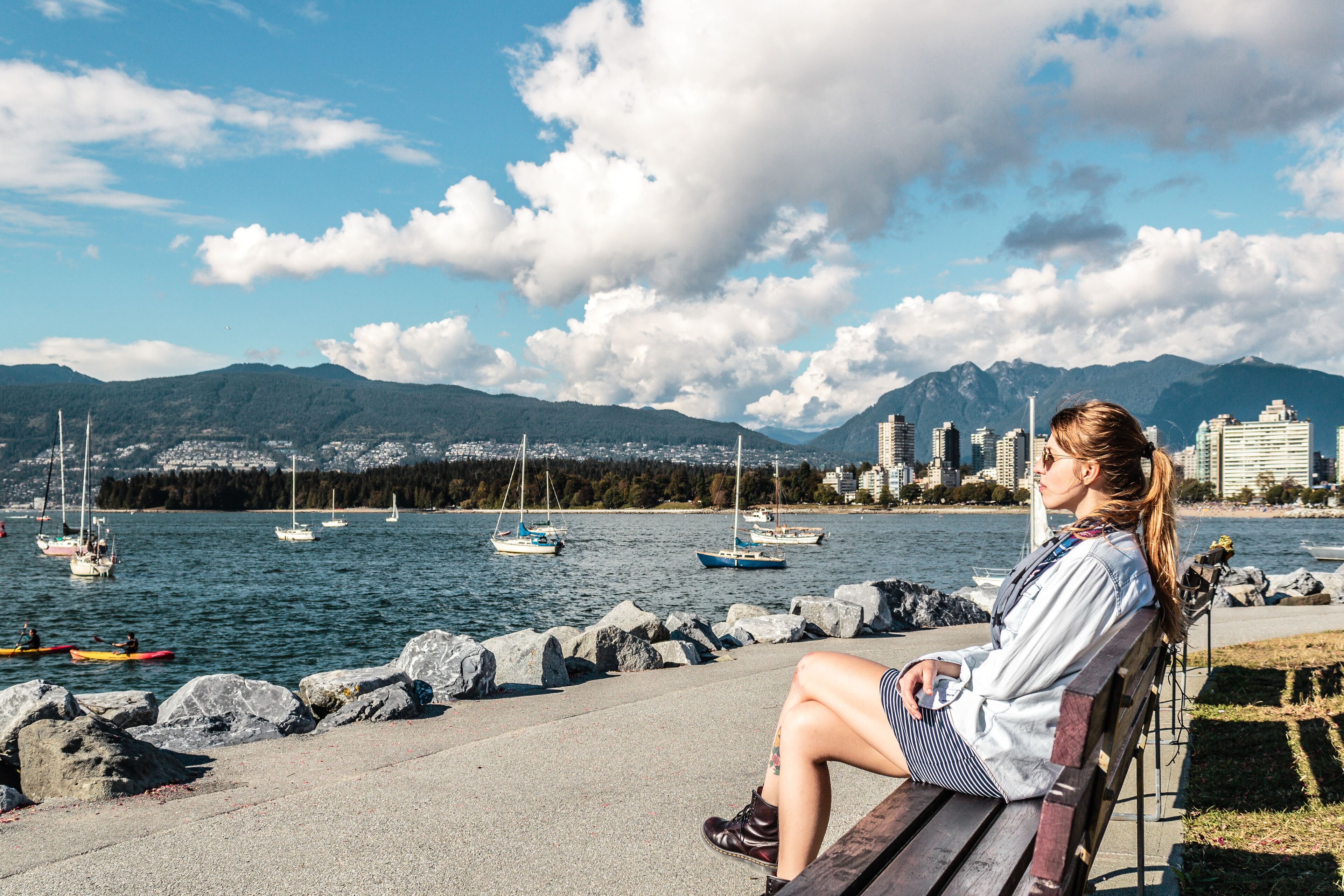 A person relaxes on a bench by the sea, gazing at sailboats and mountains under a partly cloudy sky.