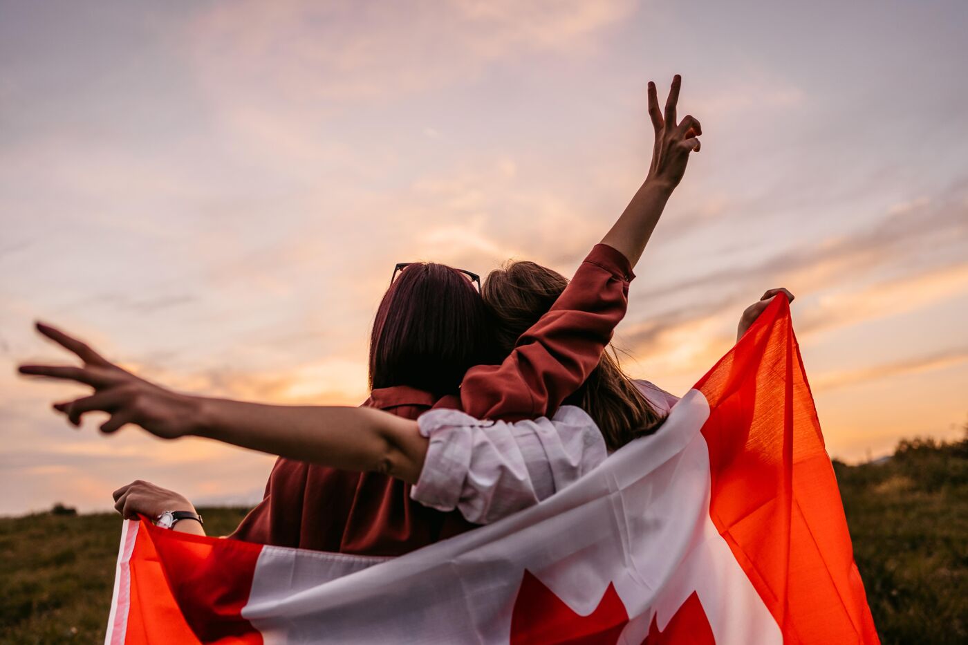 Two friends draped in a Canadian flag celebrate outdoors, raising their arms in a gesture of joy and peace. The warm sunset sky enhances the scene’s sense of camaraderie and national pride.