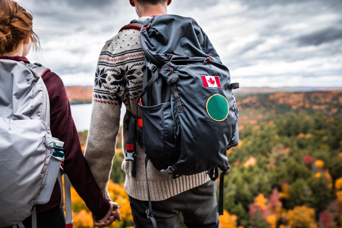 A couple holds hands while hiking, facing a scenic autumn landscape. The man carries a backpack adorned with a Canadian flag, symbolizing national pride. Both wear warm clothing suitable for the cool, clou