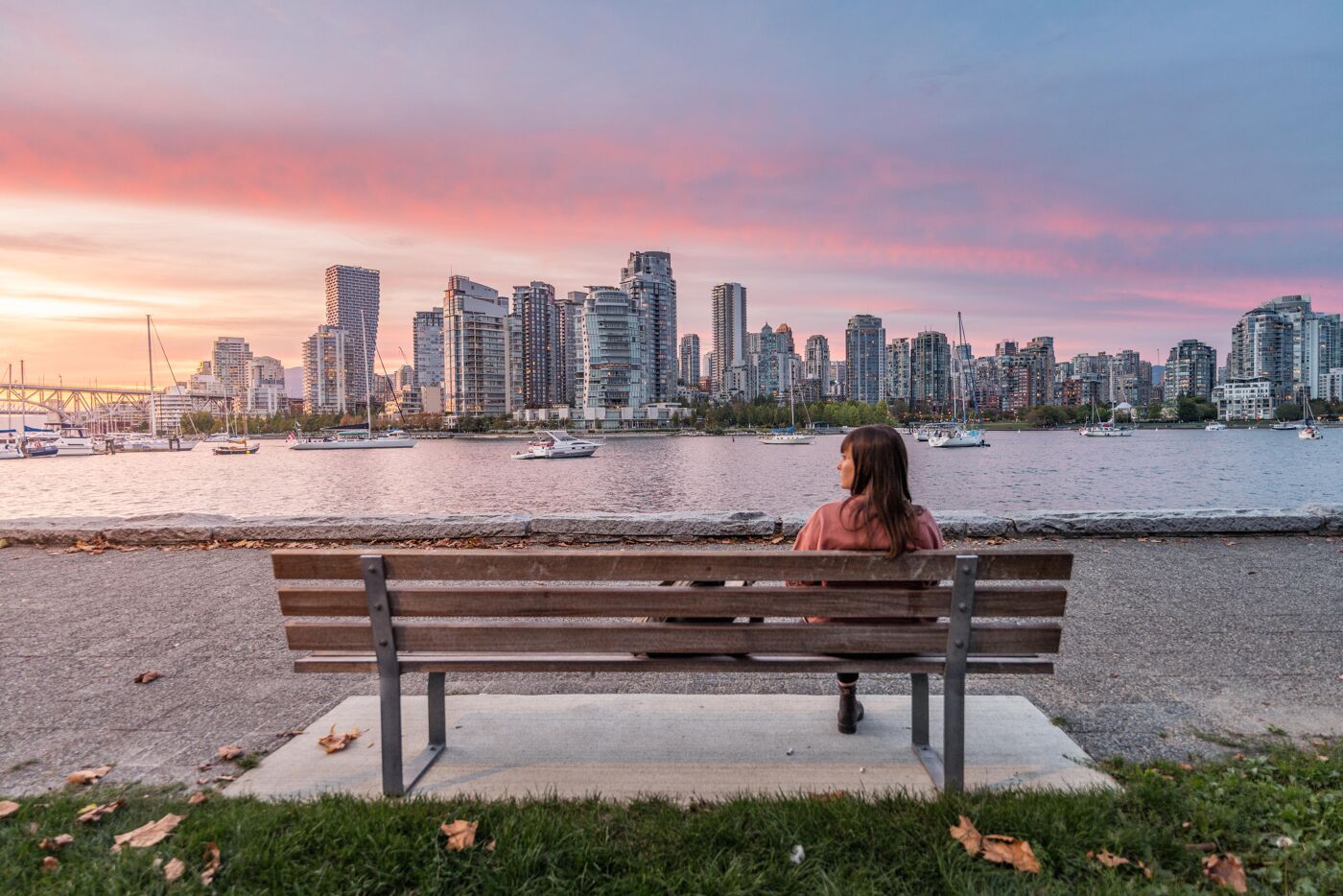 A woman sits on a bench overlooking the Vancouver skyline at sunset. The sky is painted with soft pink and purple hues, reflecting off the water, creating a serene and contemplative atmosphere.

