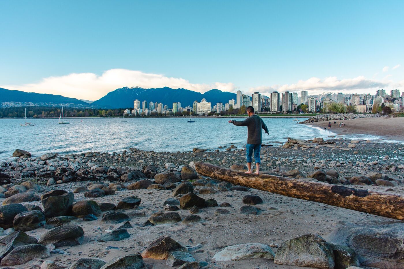 A man balances on a large driftwood log along a rocky beach in Vancouver, with the city skyline and mountains in the background. The scene captures the blend of urban and natural beauty on a calm, clear day.