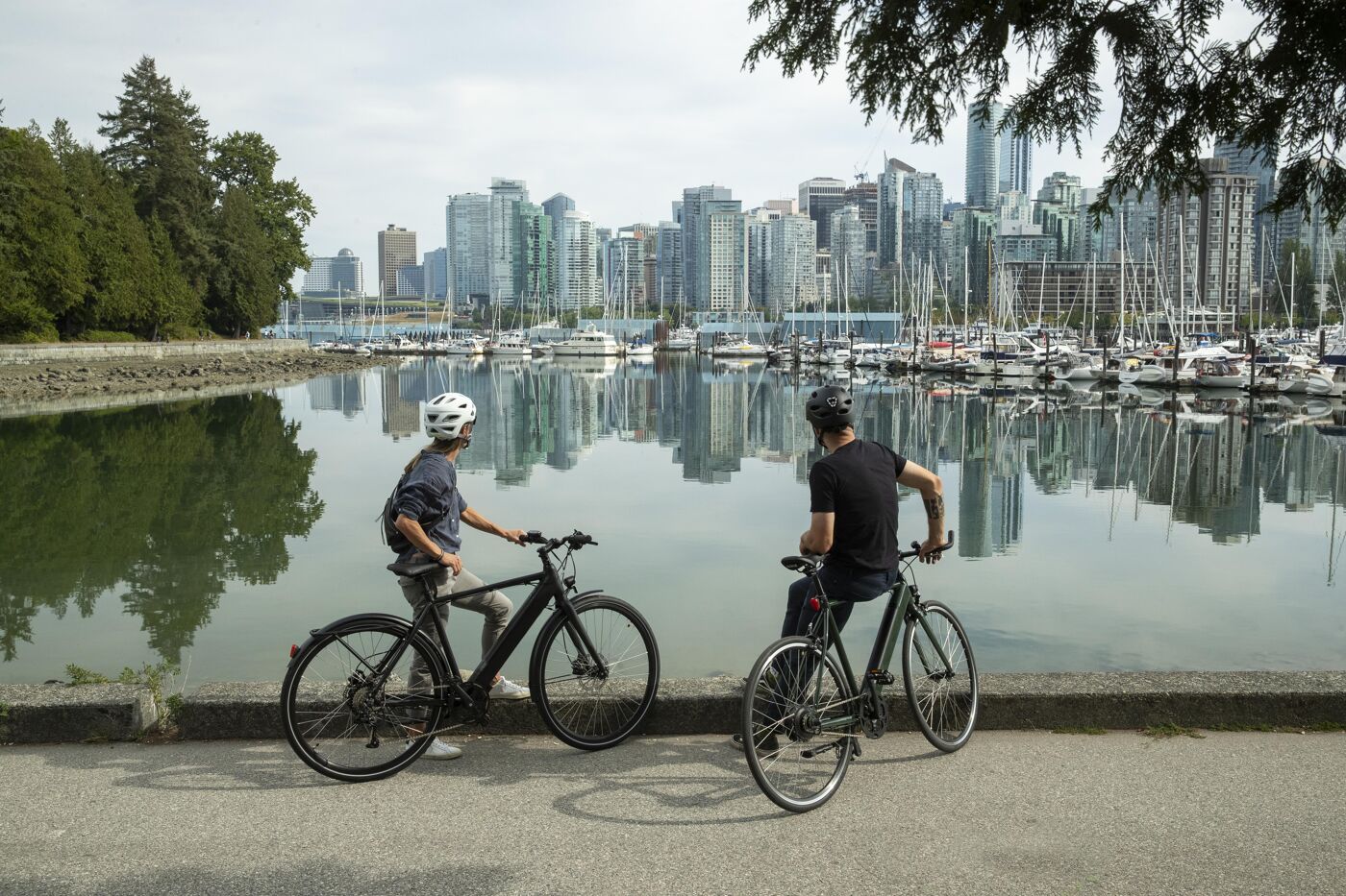 Two cyclists pause to take in the scenic view of Vancouver's waterfront, with boats docked and the city skyline reflecting on the calm water. The mix of urban landscape and nature creates a peaceful atmosphere.