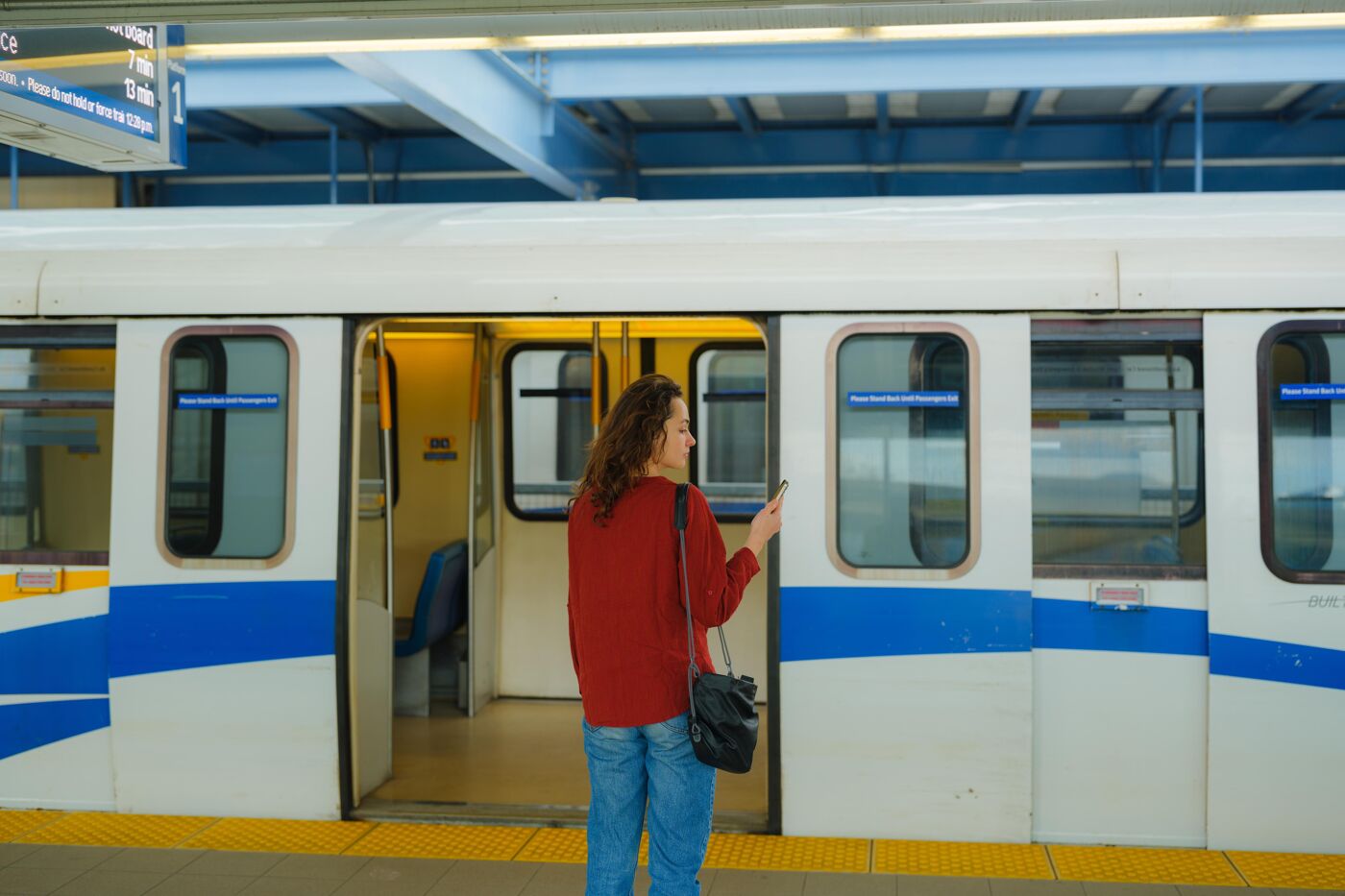 A woman stands on a train platform, looking at her phone as she waits to board the train. The train doors are open, revealing the interior of the train car. The train is white with blue accents.
