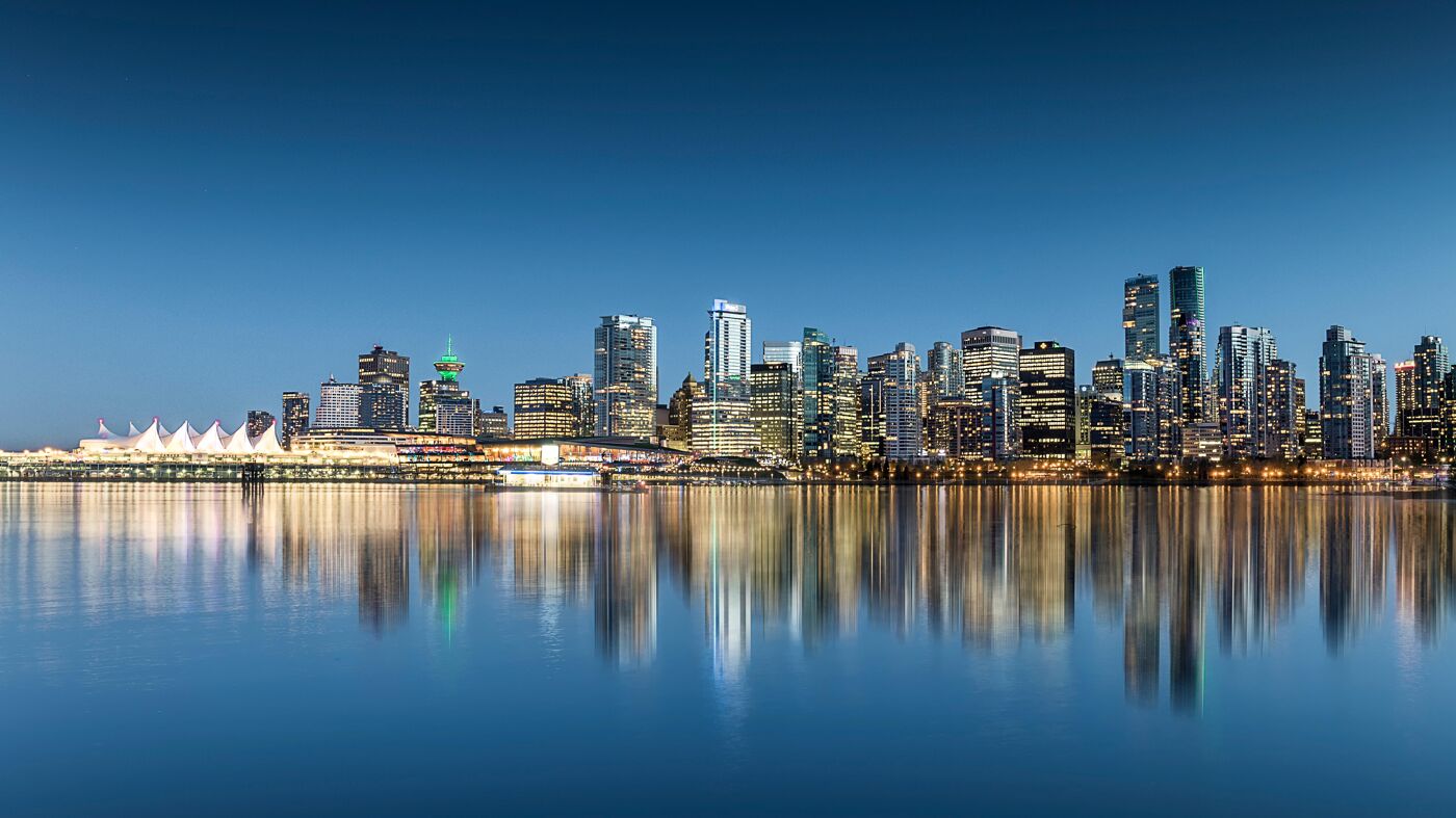 A panoramic view of the Vancouver skyline featuring illuminated skyscrapers reflected in calm waters under a clear sky. The Vancouver Convention Centre is visible on the left.