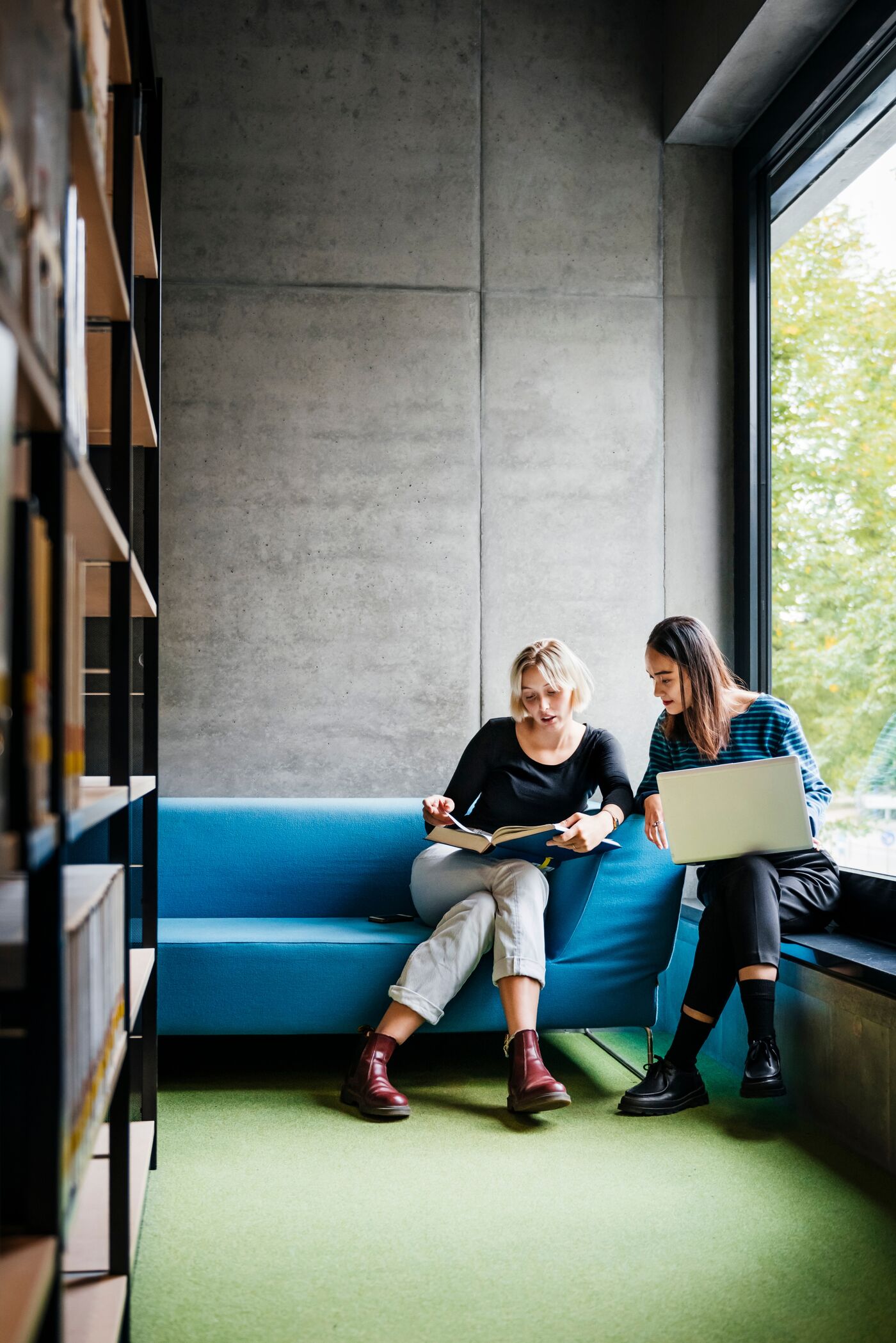 Two friends sitting on a couch and working together using a laptop and a book in a quiet library space.