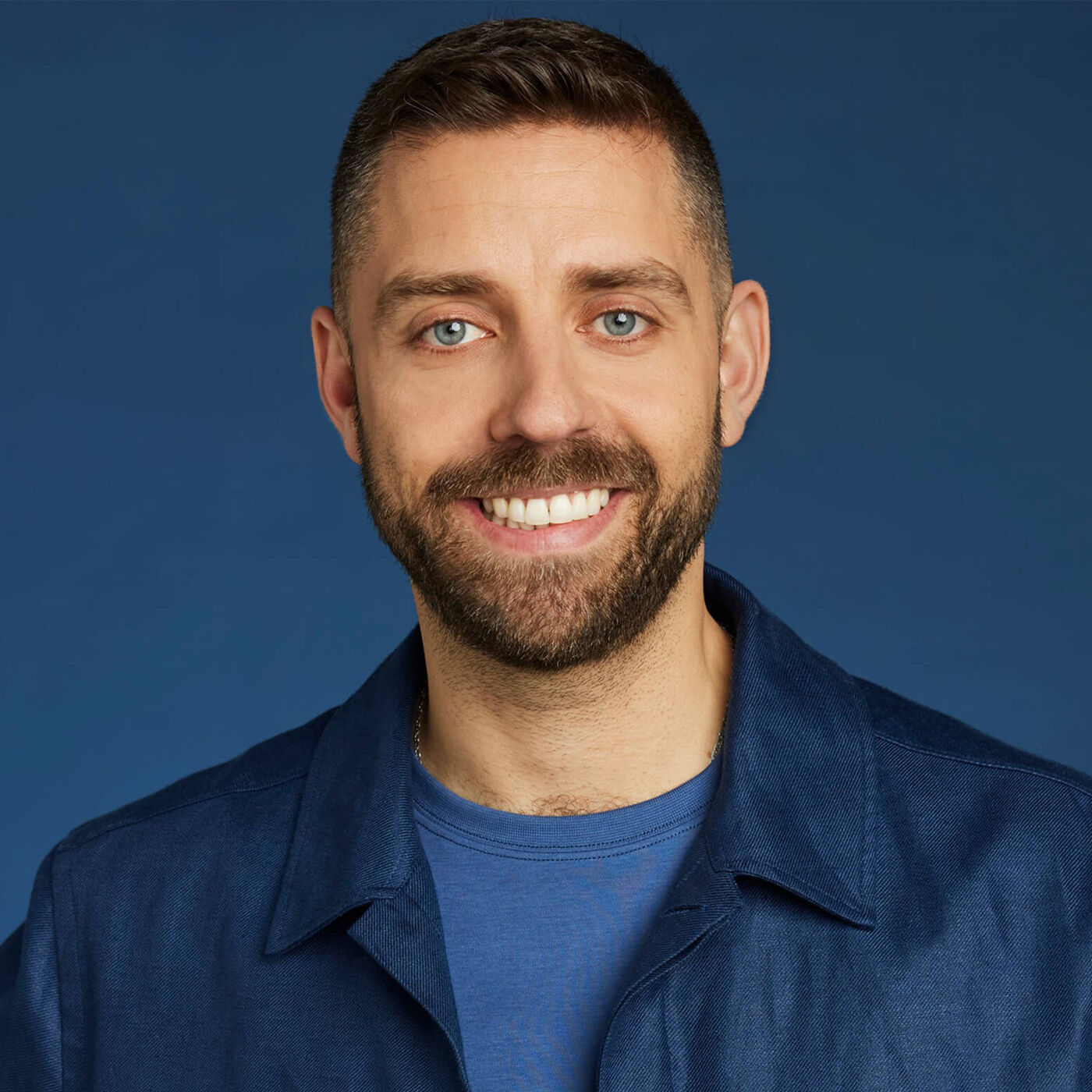 Close-up portrait of a smiling man with short brown hair and a beard, wearing a dark blue collared shirt.