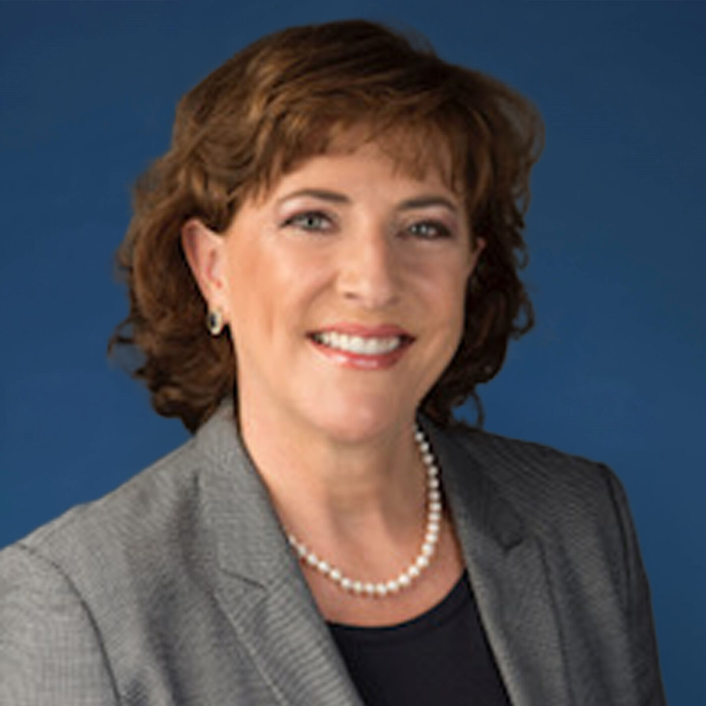 Close-up shot of a woman with shoulder-length brown hair wearing a gray blazer and a pearl necklace against a blue background.