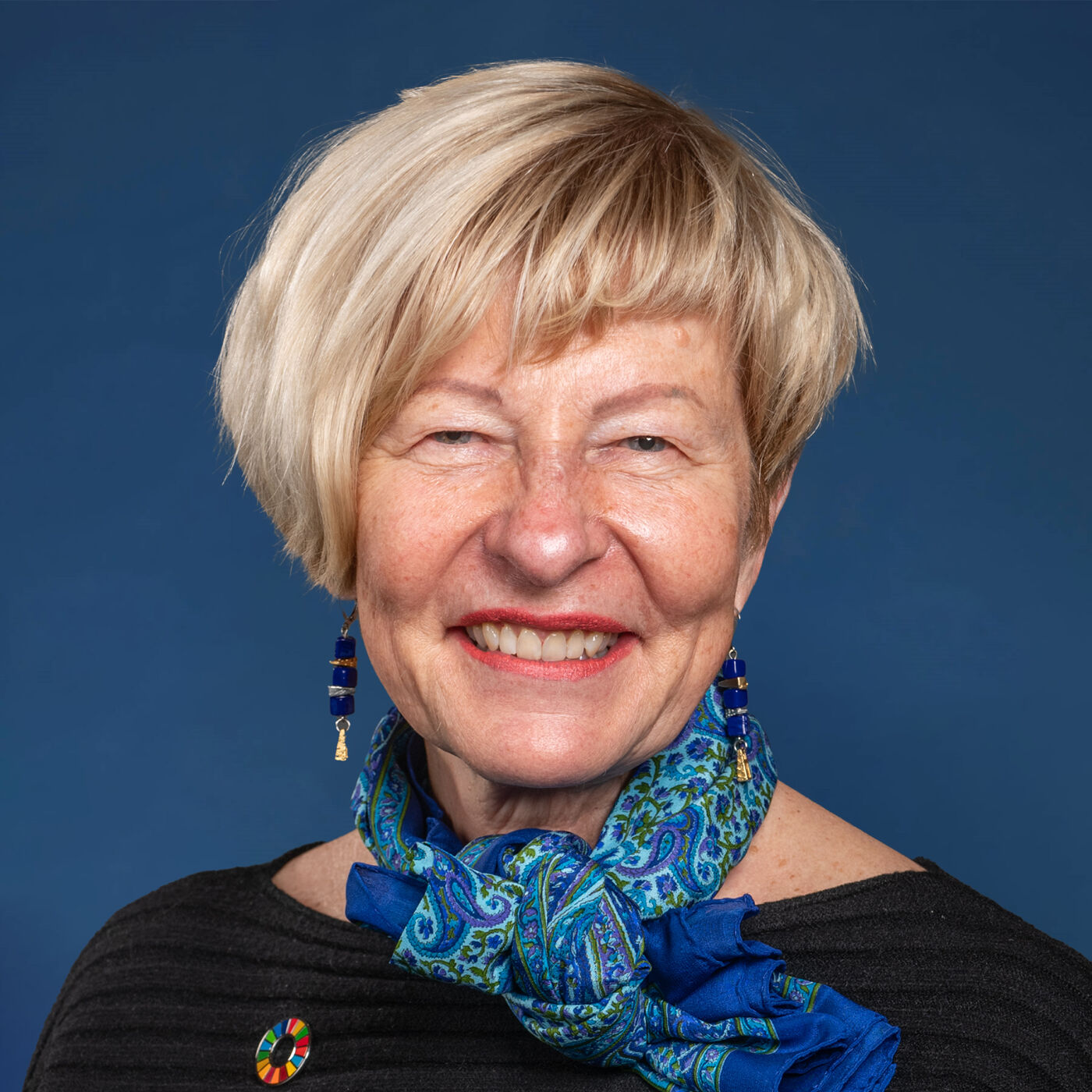 Close-up portrait of a smiling woman with short blonde hair, wearing a blue paisley scarf and earrings.