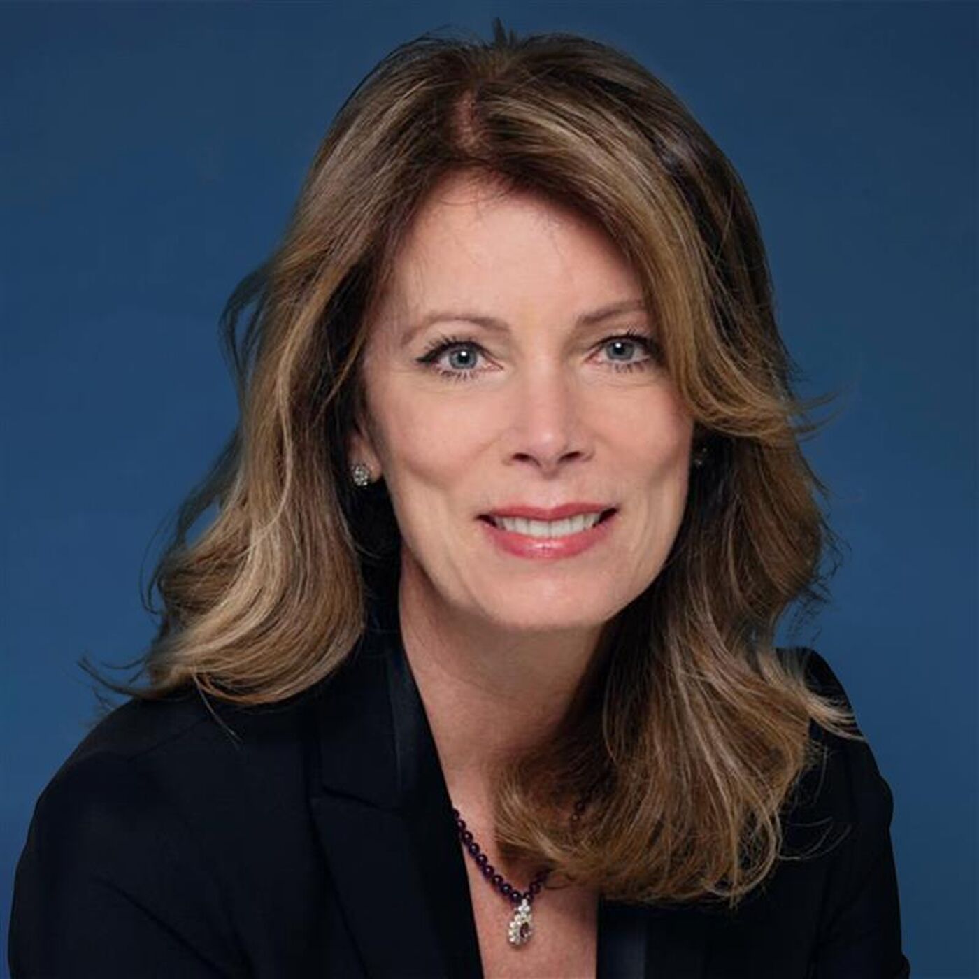 Close-up portrait of a woman with shoulder-length brown hair, wearing a black blazer and a dark-colored necklace, against a blue background.
