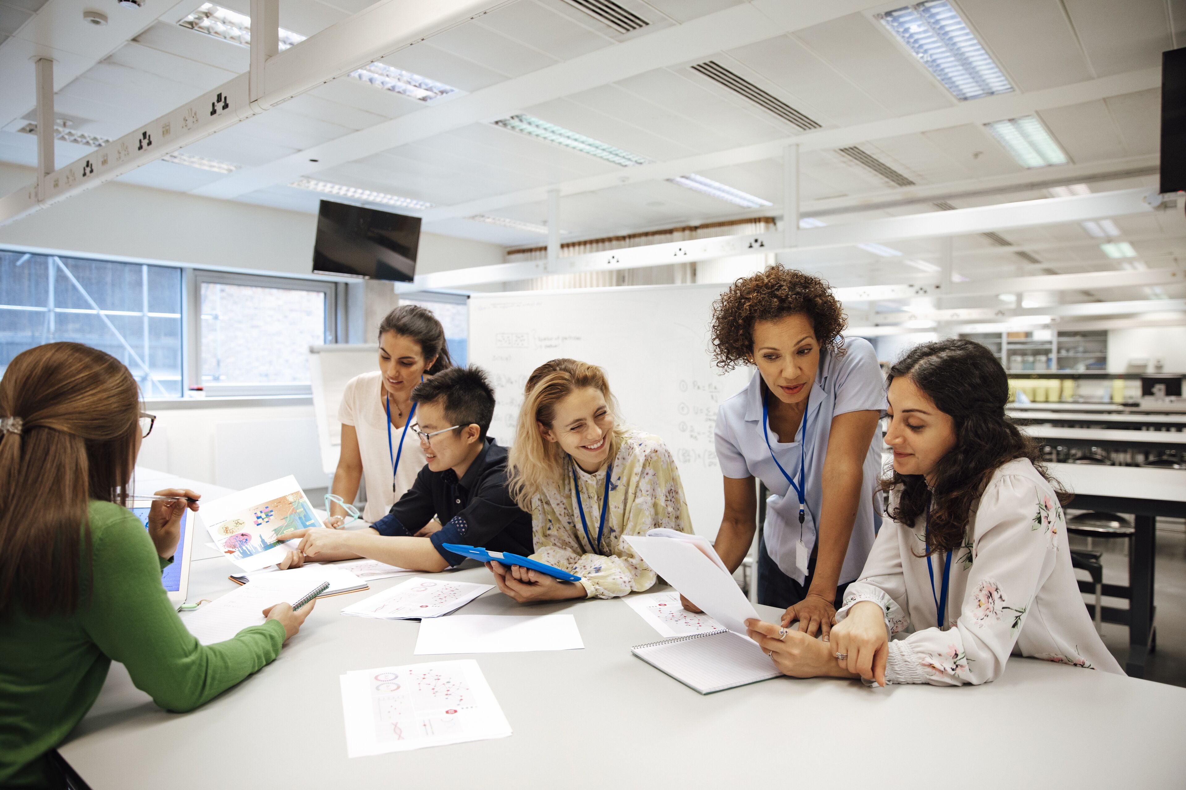 Teacher with a group of university students, in a laboratory classroom. The instructor is considering one of the students work, the mood is light hearted and positive. Other classmates are discussing things with each other. This is a realistic teaching scenario, with candid expressions. This is a multi-ethnic group of women. In the background there is a white board with mathematical formula written on it. All ladies are wearing id tags.