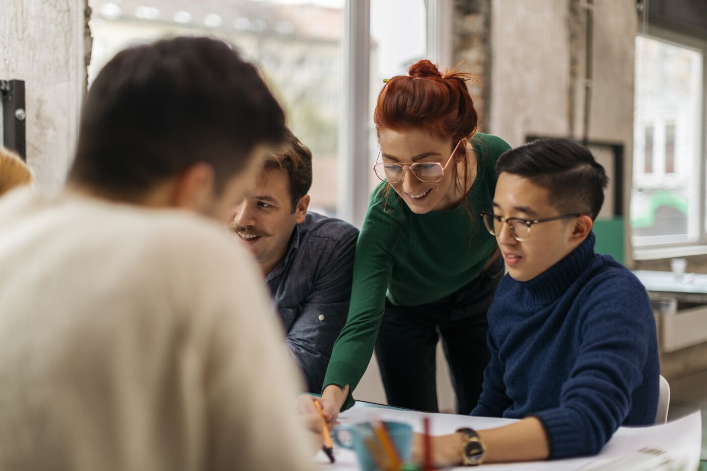 Young international team of colleagues in a design studio working on a paper sheets. Discussing plans and making forecast. Confident red hair girl explaining plans to others.