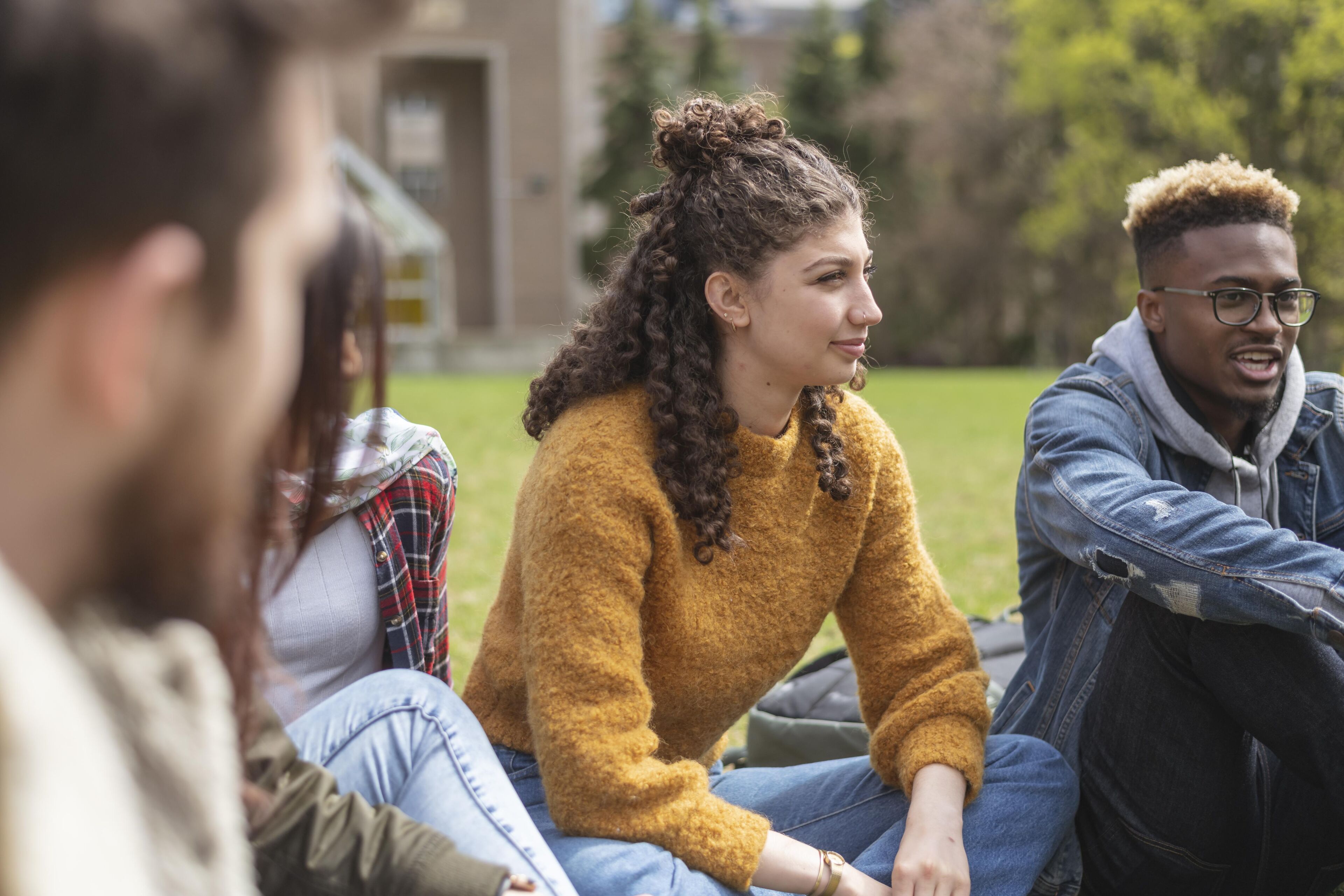 A group of university friends spends time outside on their campus. They are smiling while relaxing. They are all dressed casually, yet fashionable.