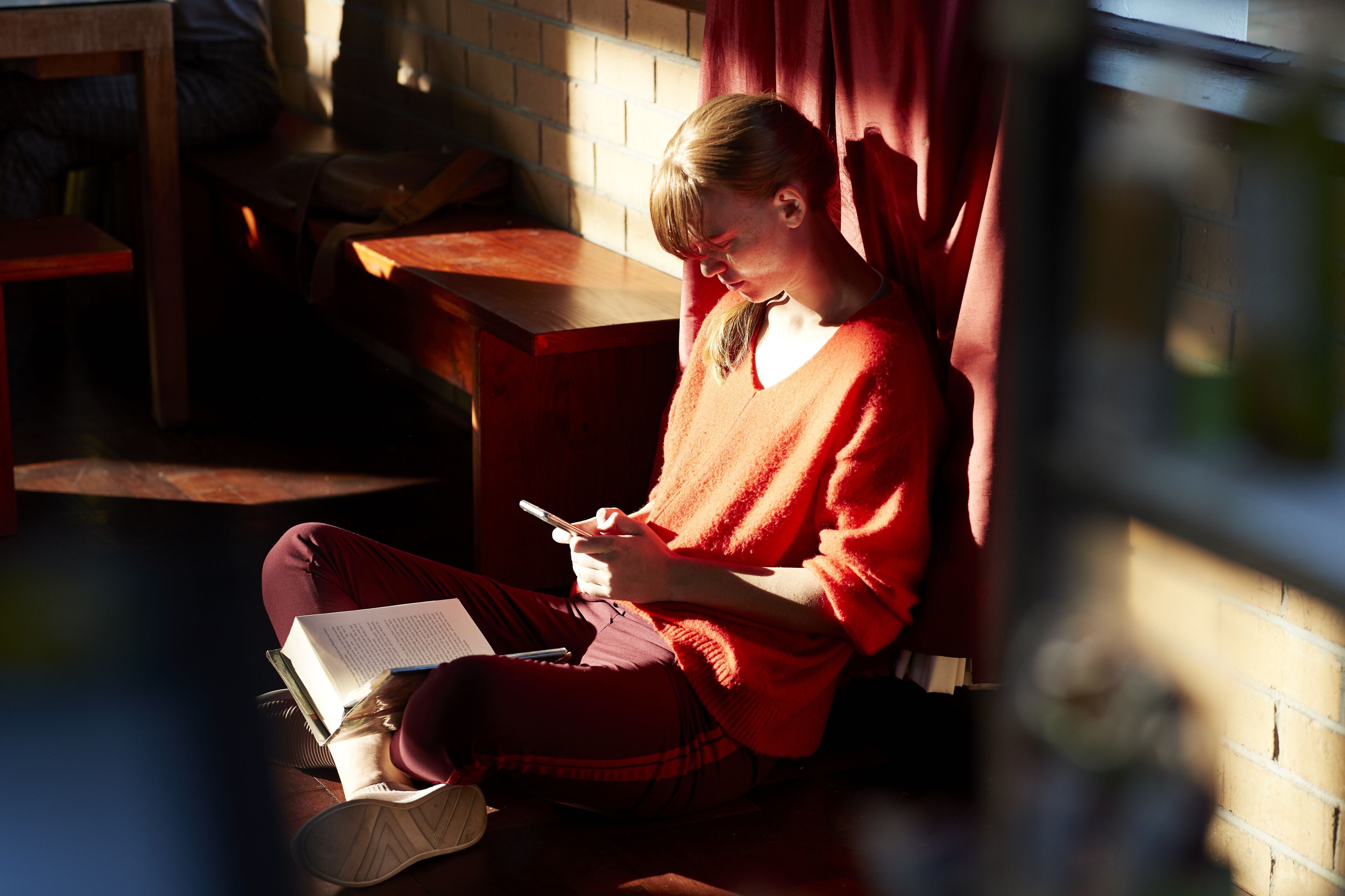 Full length of young woman leaning on wall while sitting with book and using smart phone on floor at cafeteria in university