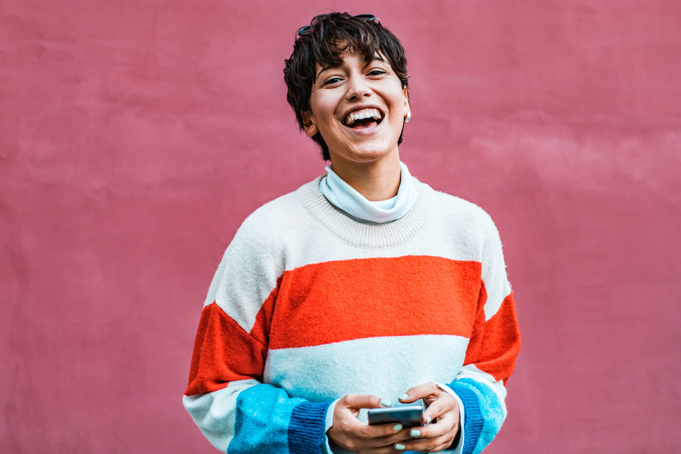 A young woman with short, dark hair smiles broadly while holding a smartphone in front of a pink wall, wearing a horizontally striped sweater with white, orange, and light blue sections.