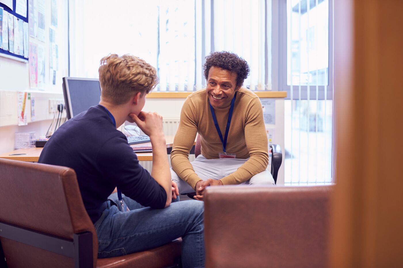 Two men are sitting in chairs, engaged in a conversation, in what appears to be an office or counseling setting.