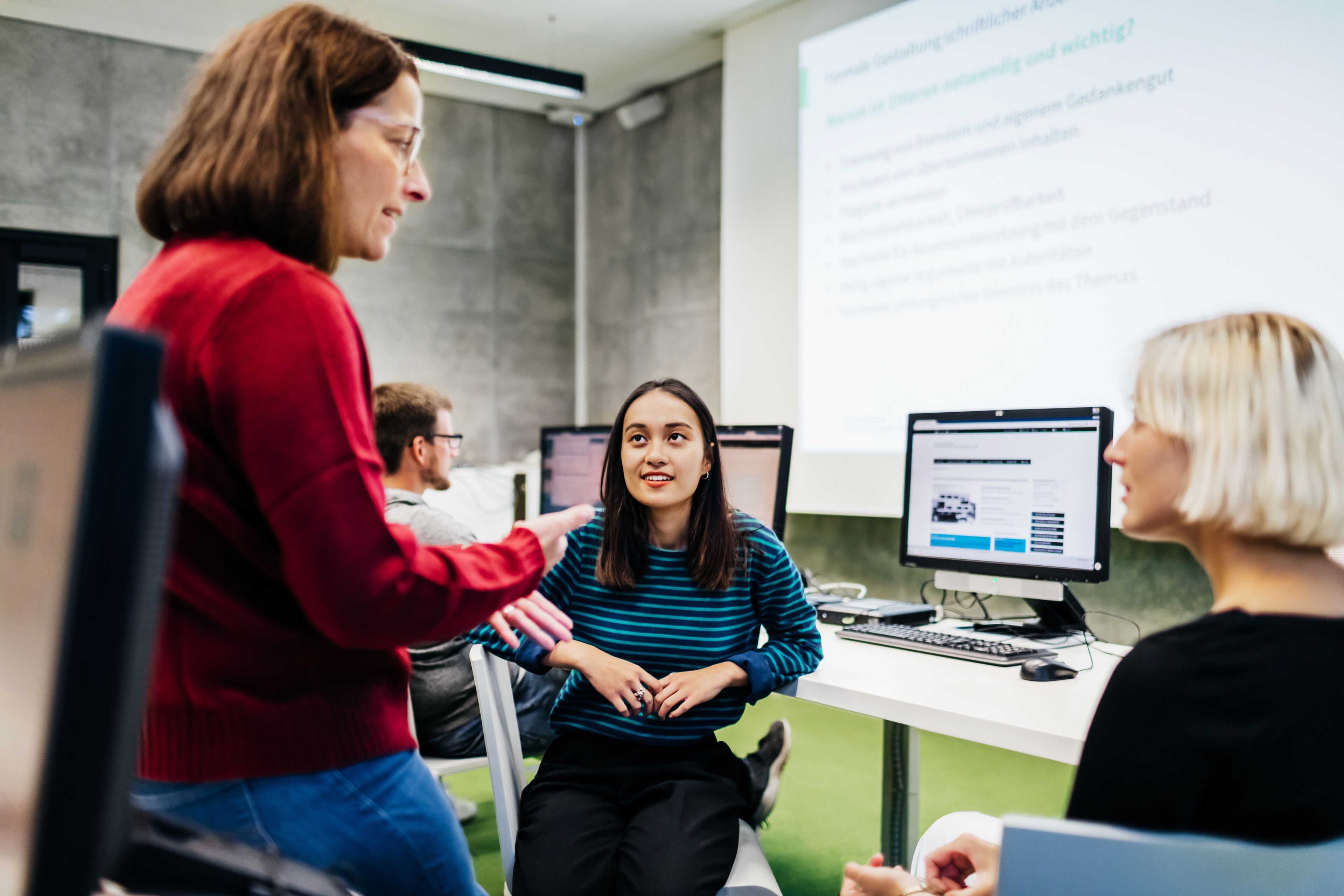 A tutor leaning on a desk and explaining concepts to a group of her students during a seminar at university.