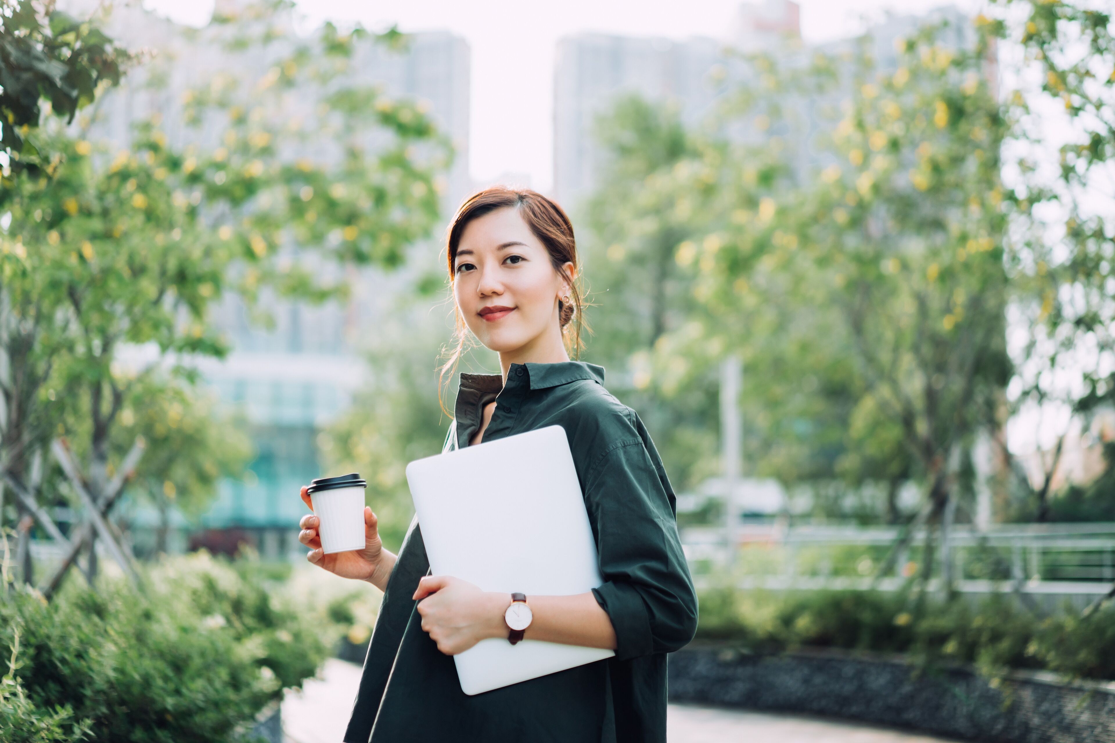 A confident young woman with a laptop and coffee, set against a lush park and modern cityscape, embodies professionalism and urban lifestyle.