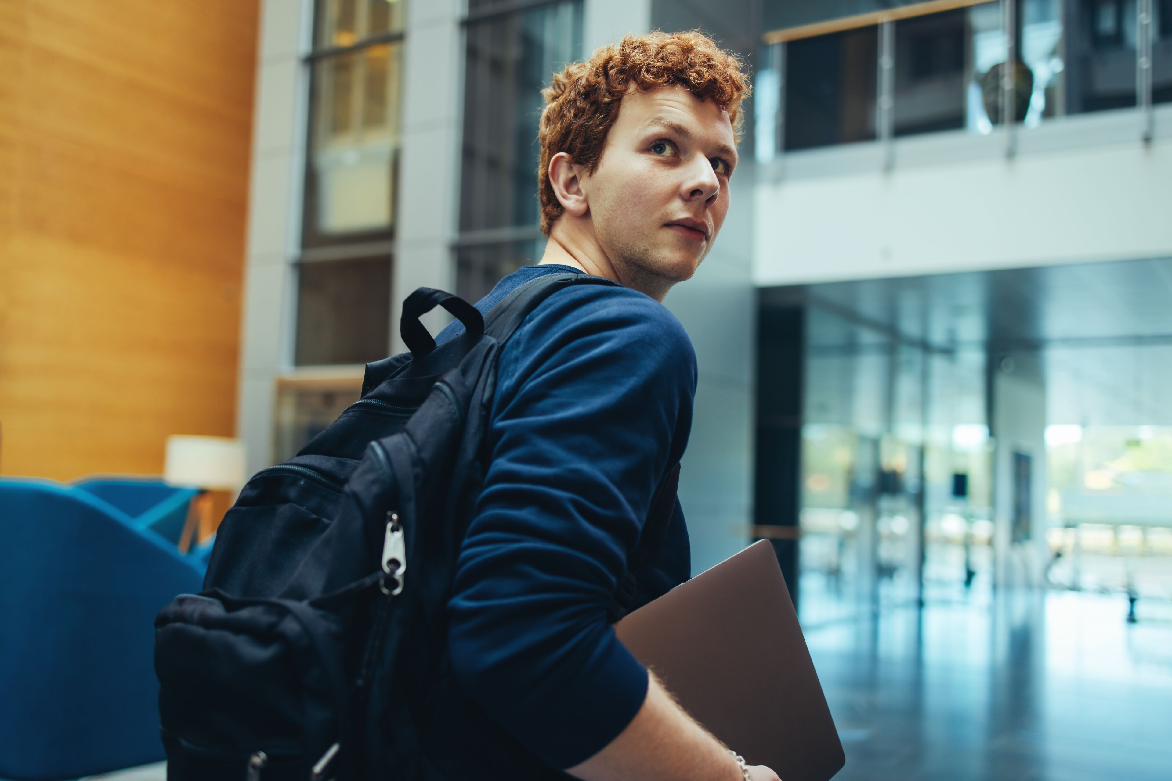 A pensive young man with curly hair, carrying a backpack and laptop, gazes out in a contemporary building.