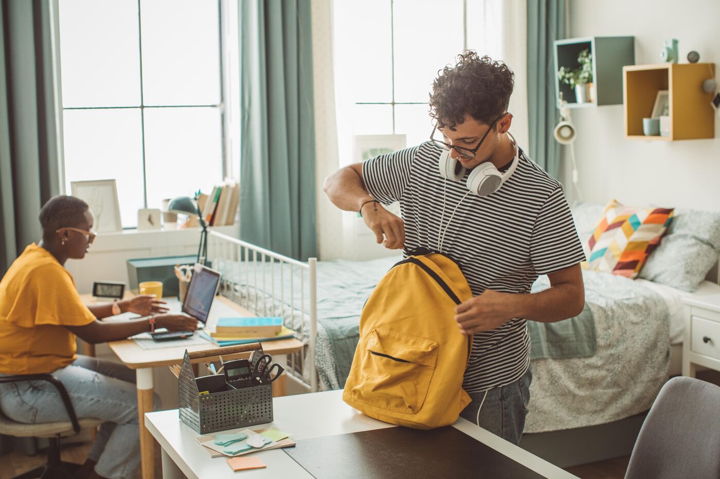 A young man packs a yellow backpack while a young woman works on a laptop at a desk in a shared bedroom.