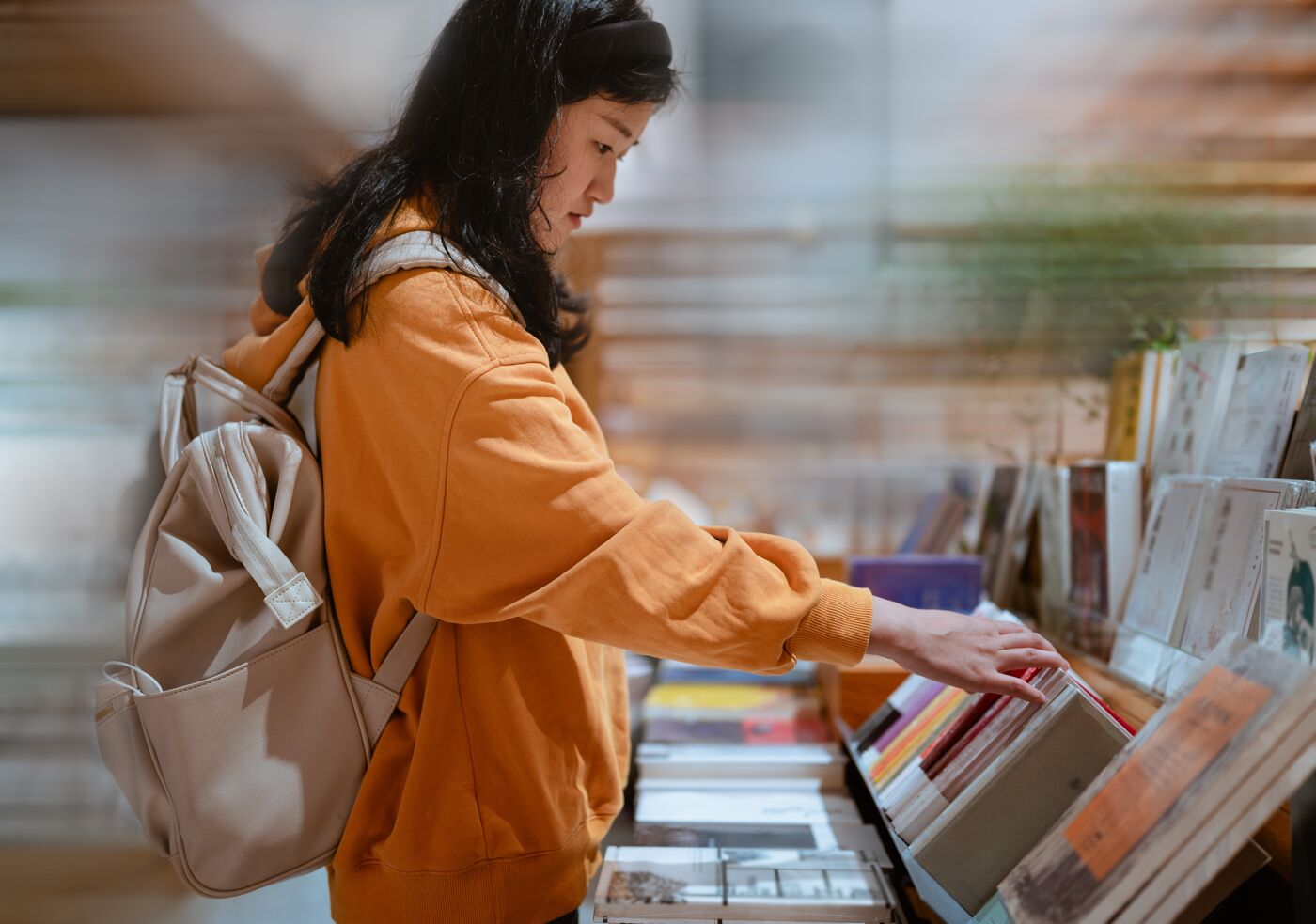 In the library, an Asian woman with a backpack selects books from the shelves