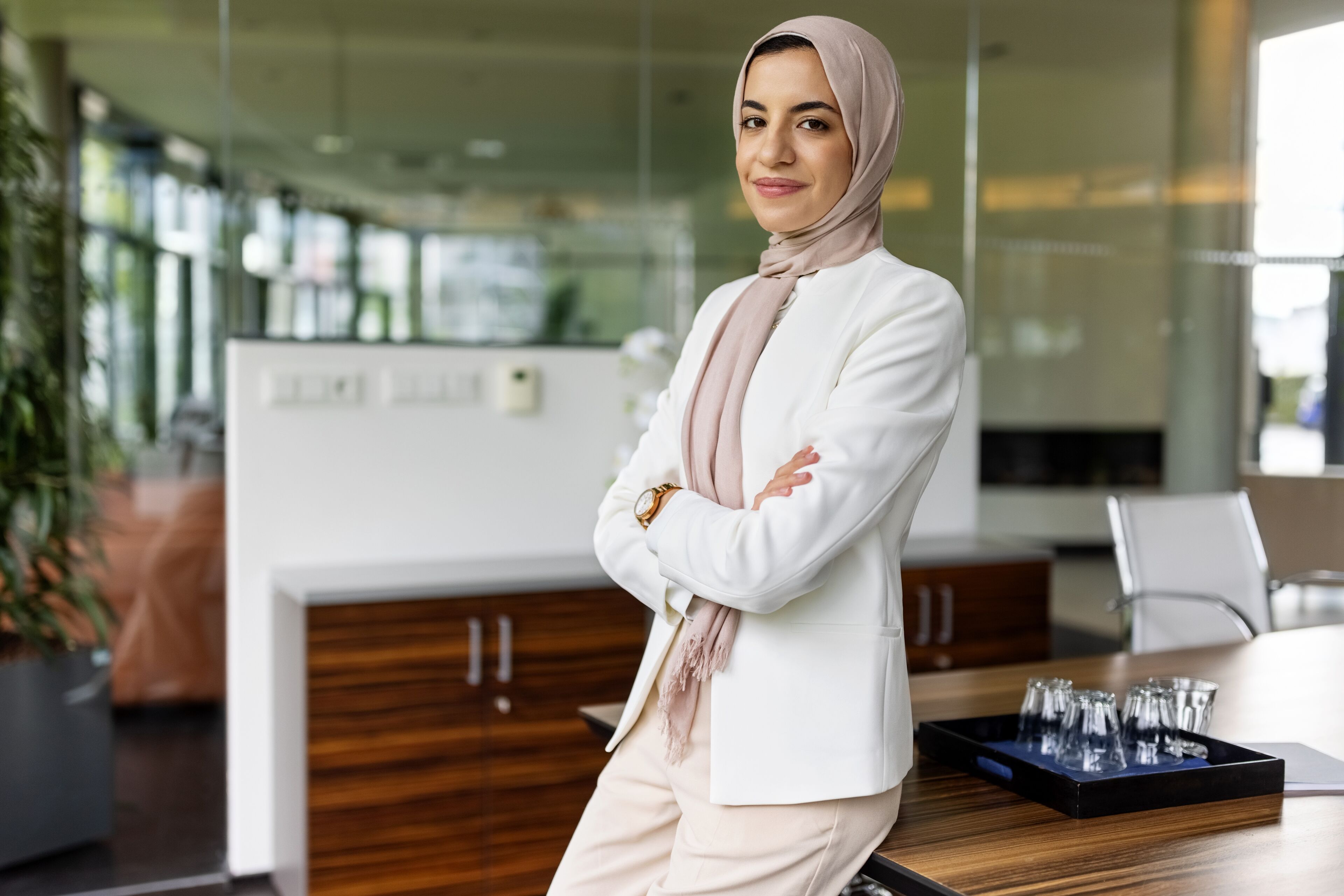Une femme posée portant un hijab se tient les bras croisés dans un bureau moderne, dégageant confiance et professionnalisme.