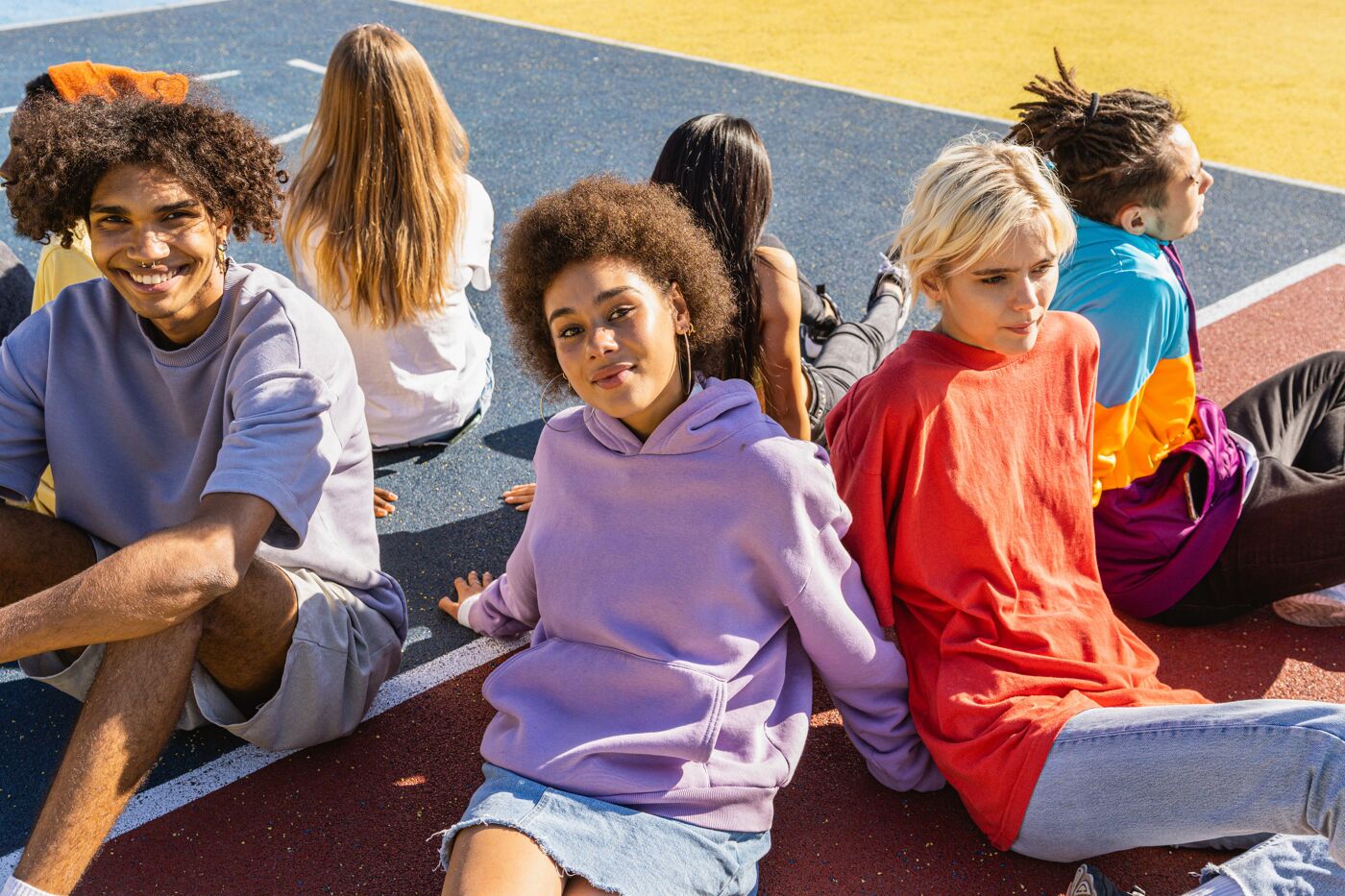 A group of six young people are sitting on a colorful court, posing for a photo. They are dressed in casual clothing, and appear relaxed and happy. The court has sections of blue, yellow, white and red.