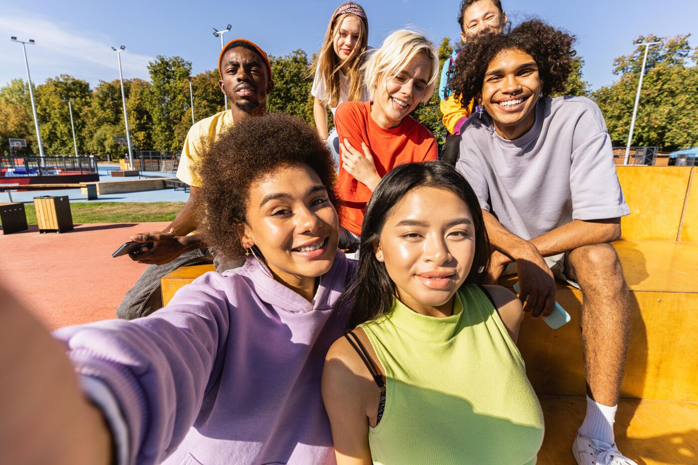 A group of young, diverse friends are posing for a selfie outdoors on a sunny day. They are smiling and appear to be enjoying each other's company in a casual setting, possibly a park or recreational area.