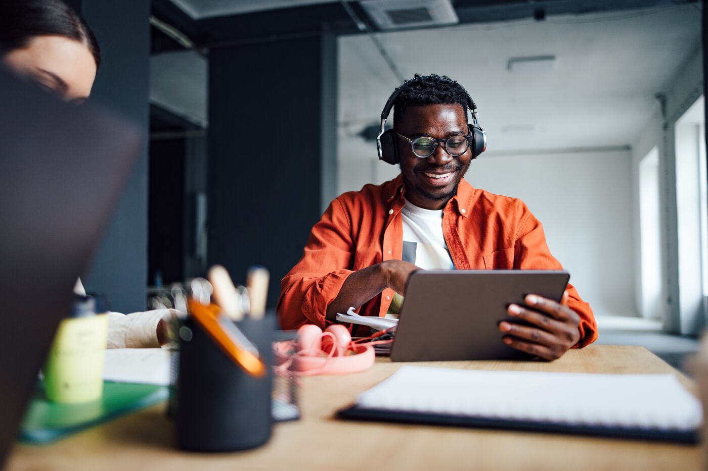 A joyful man wearing glasses and headphones interacts with a tablet, sitting at a desk with office supplies and a notebook in a bright workspace.