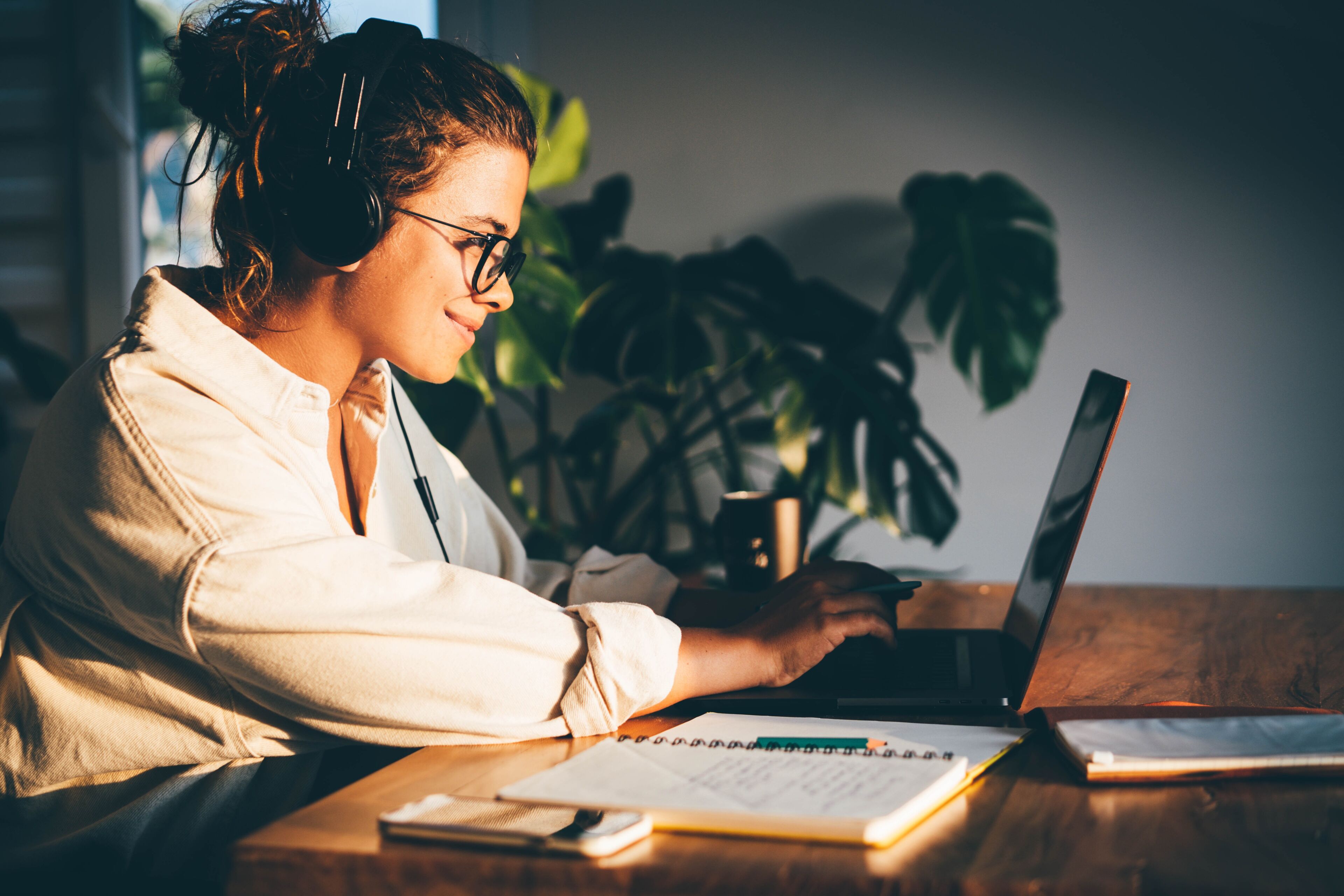 A focused woman in glasses using a laptop at a wooden desk with headphones on, notebook at hand, and a houseplant beside her, bathed in the warm glow of the sunset.