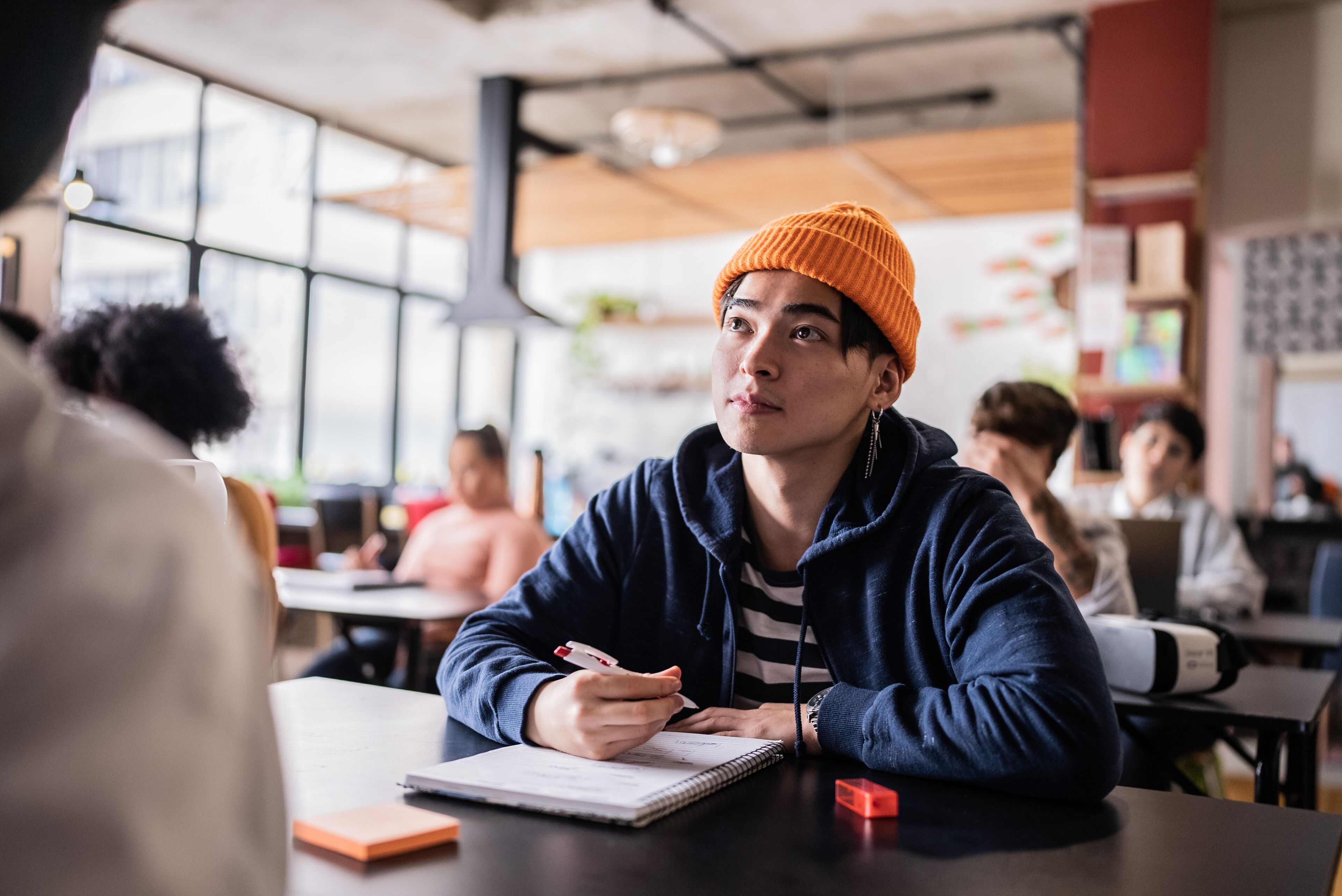  A focused young adult in a beanie and hoodie sits in a classroom setting, pen in hand, attentively listening.