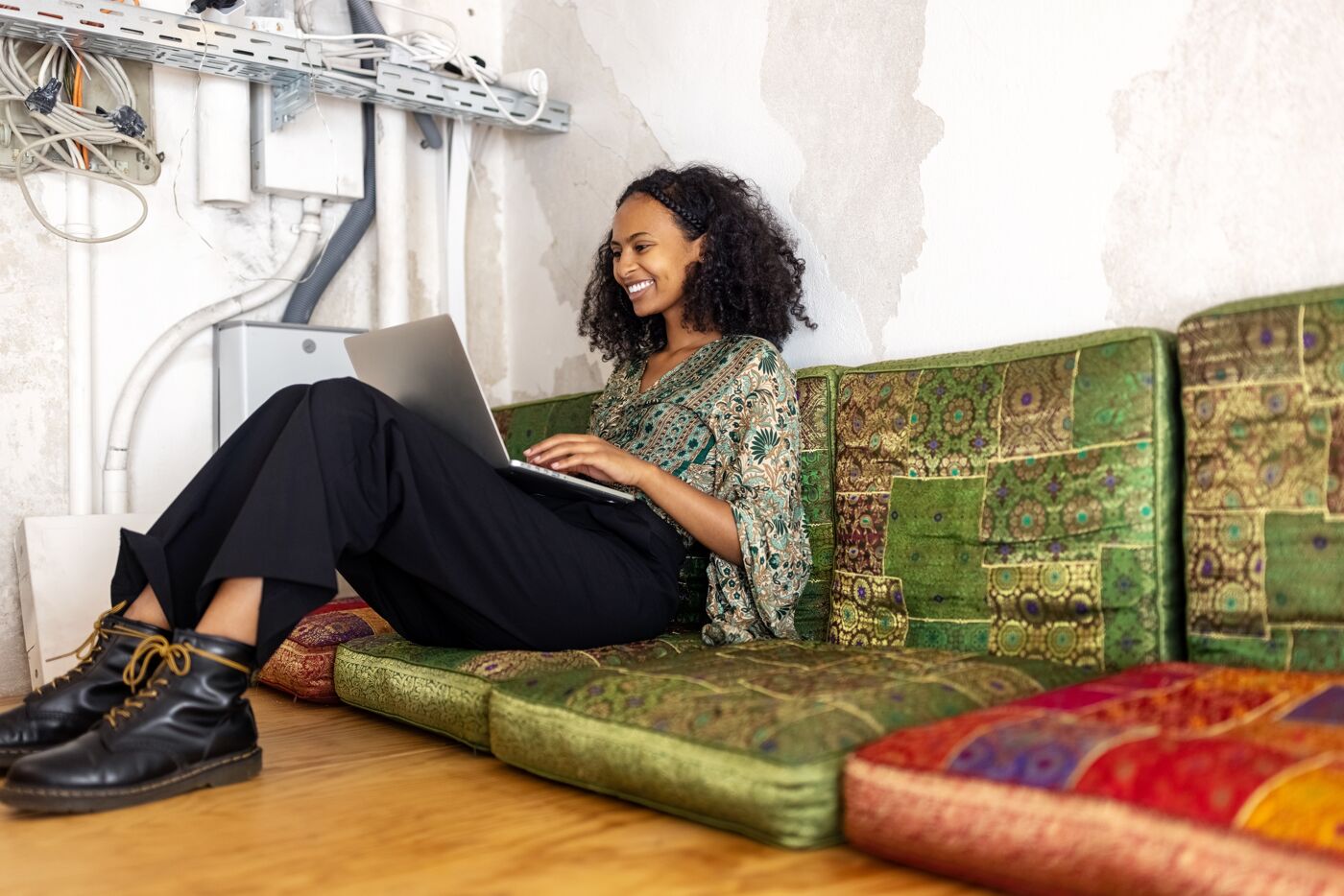 Smiling woman comfortably working on a laptop while seated on colorful cushions in an informal setting.