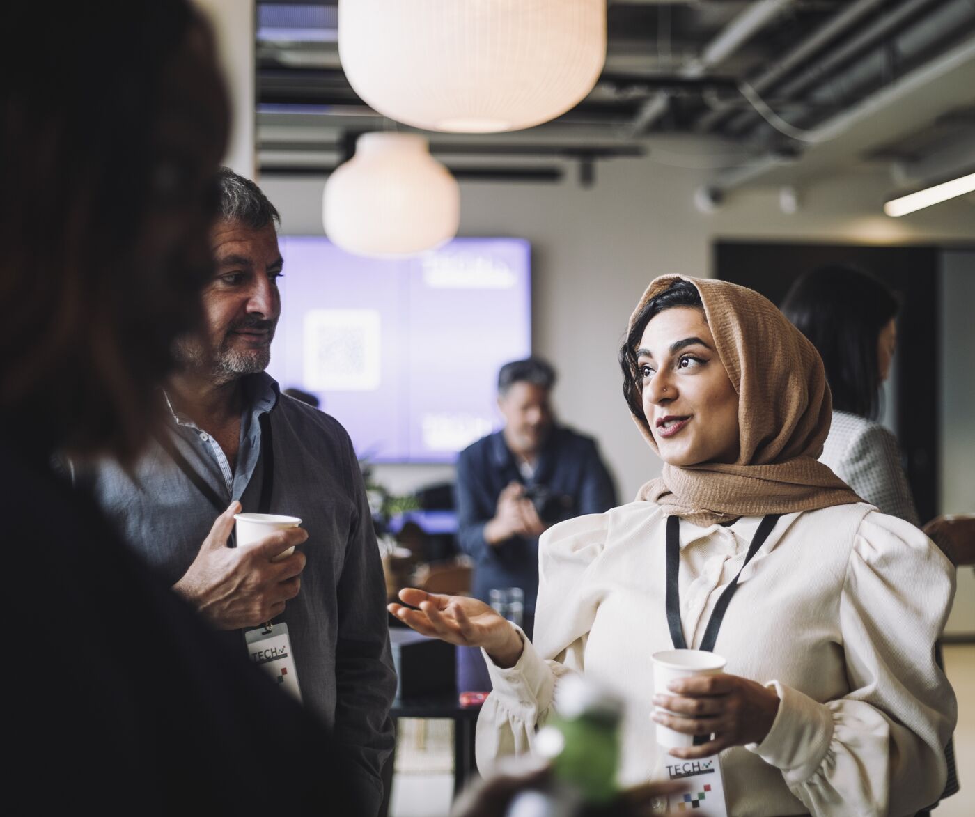 A woman in a hijab converses with colleagues over coffee in a relaxed, contemporary networking event setting.