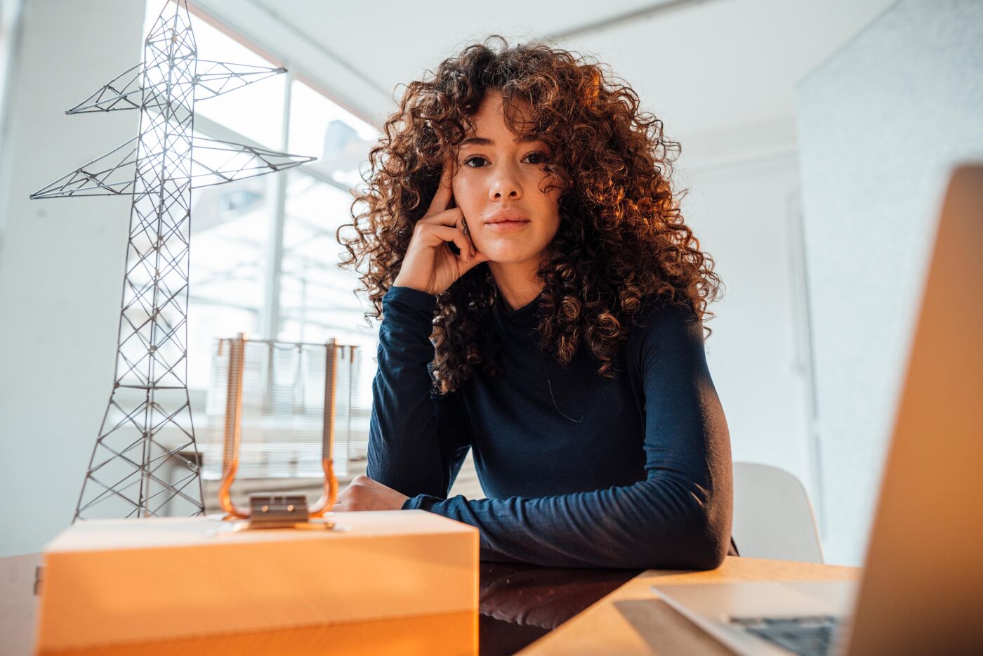 A woman with curly hair looks towards the camera, resting at a table with a model of a power line tower and a laptop beside her.